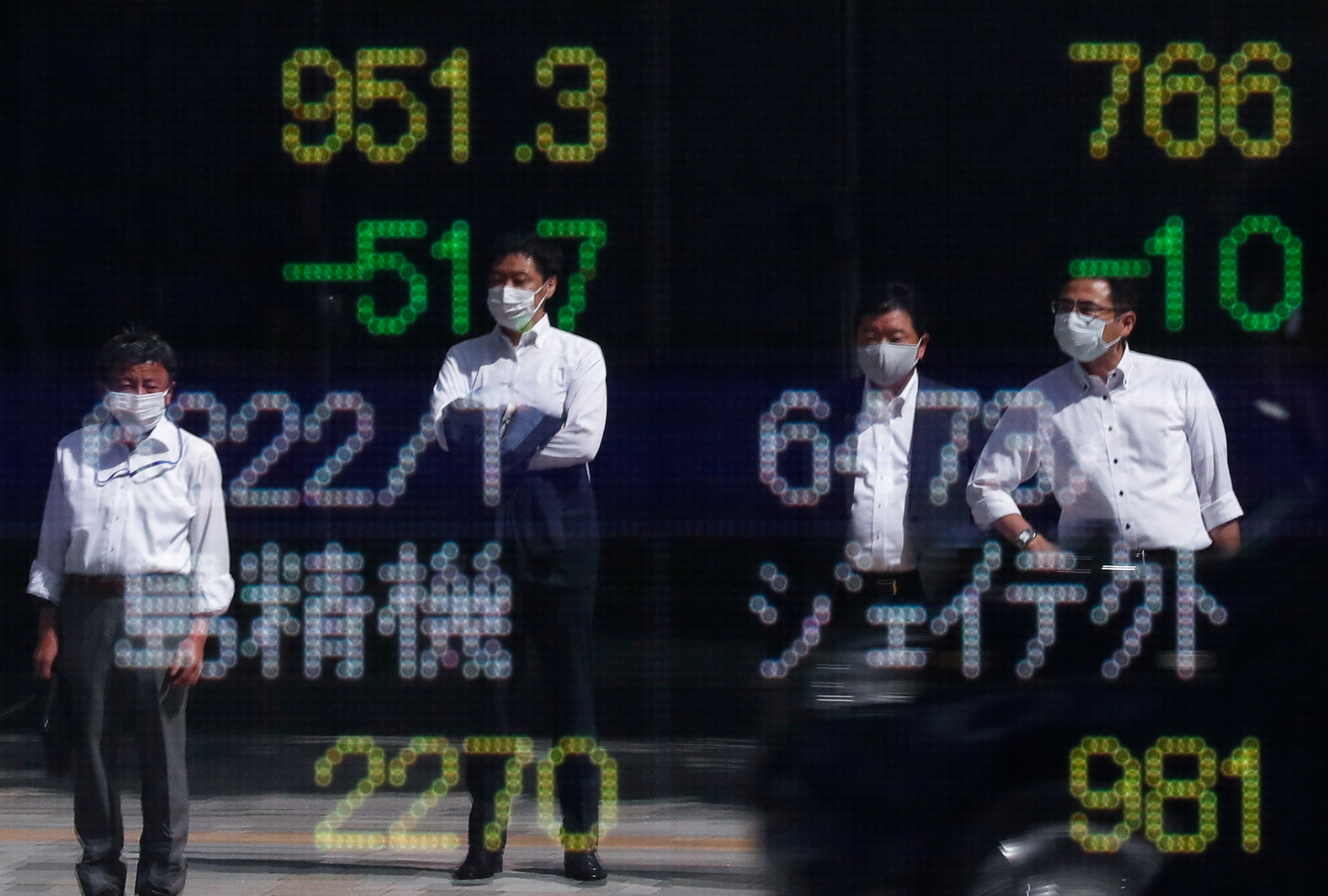 Passersby wearing protective masks are reflected on an electronic board displaying stock prices outside a brokerage amid the COVID-19 outbreak, in Tokyo