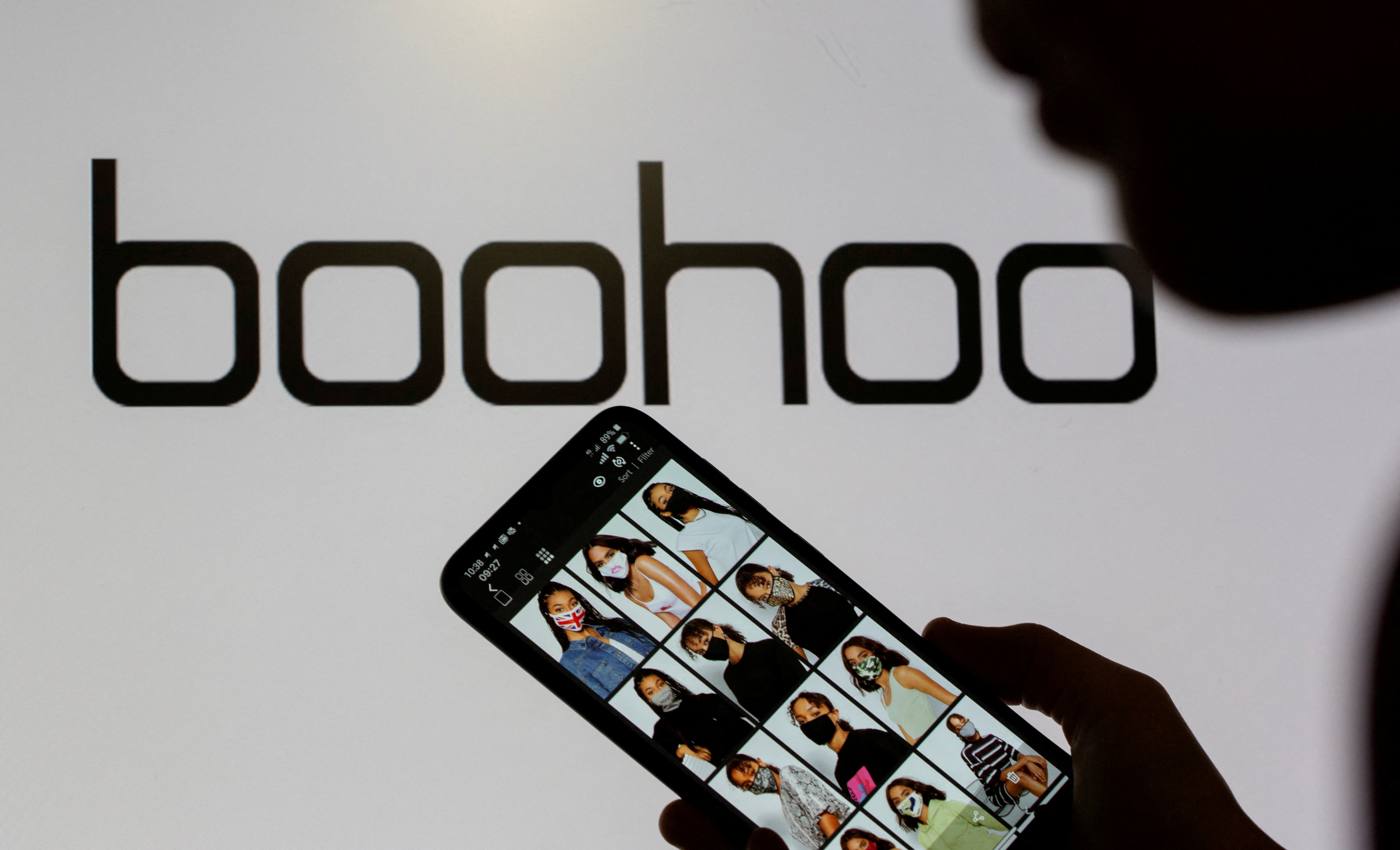 A woman poses with a smartphone showing the Boohoo app in front of the Boohoo logo on display in this illustration