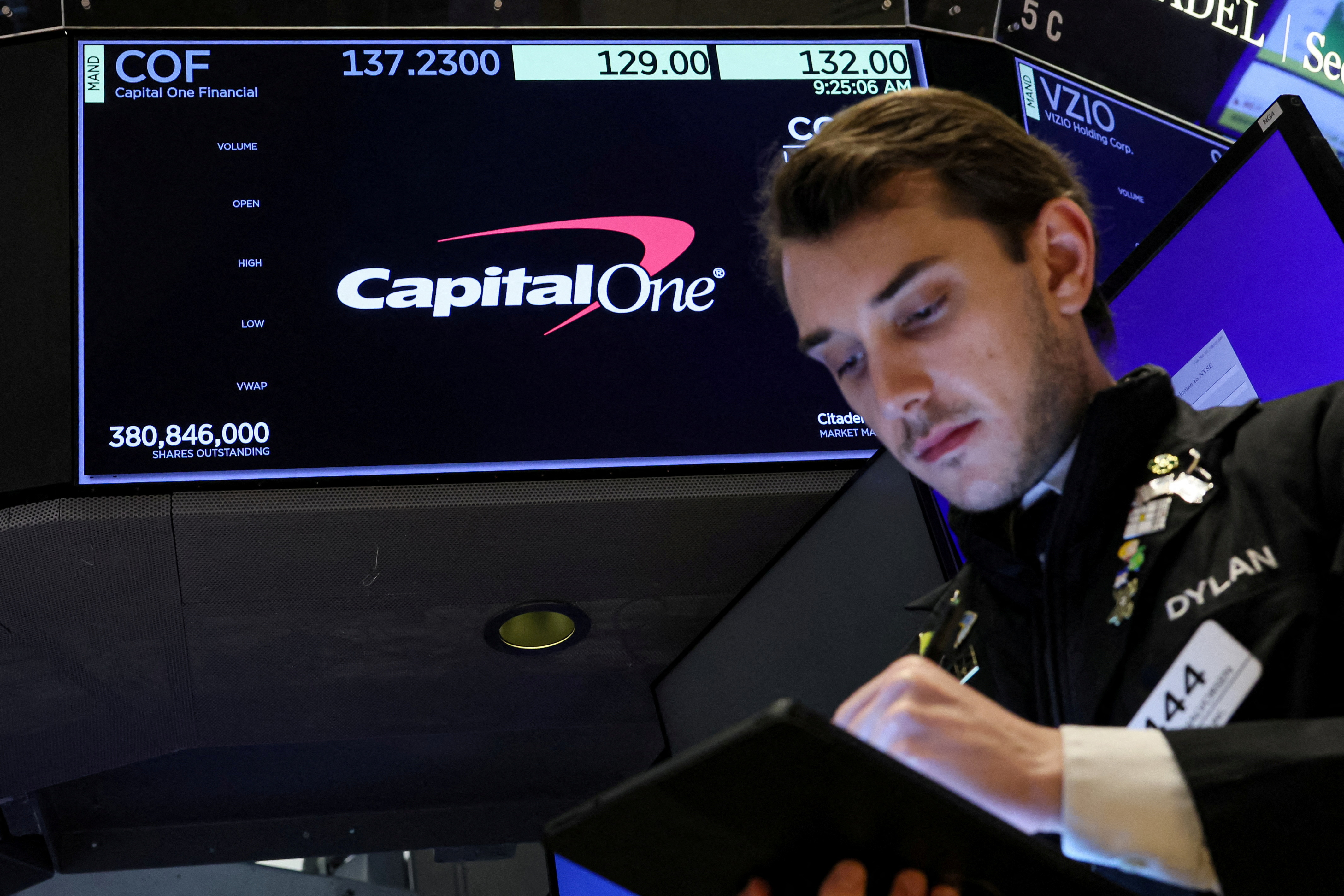A screen displays the logo and trading information for Capital One Financial as a trader works on the floor at the NYSE in New York