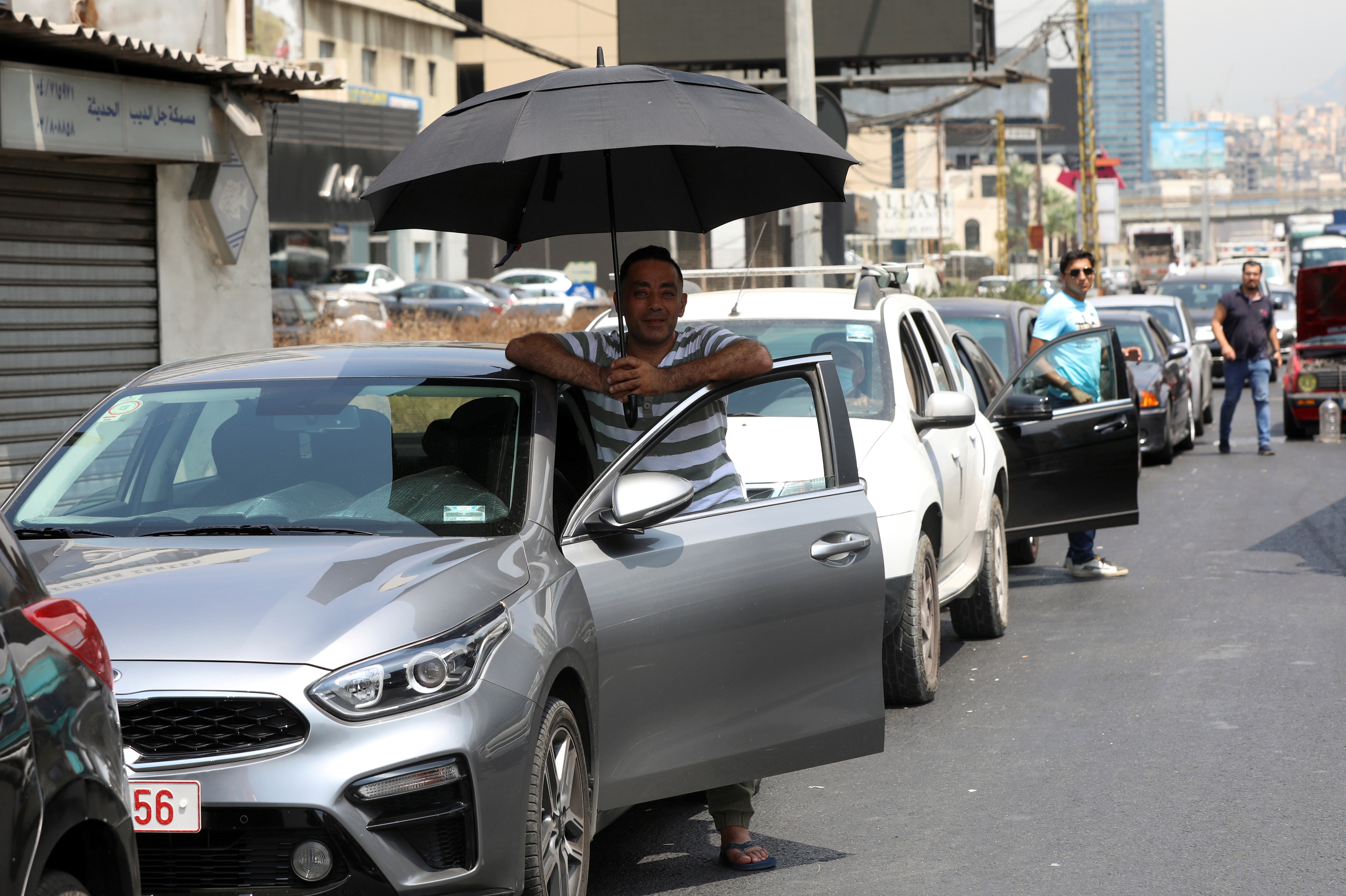 A man shields from the sun with an umbrella as he waits to get fuel from a gas station in Jal el-Dib