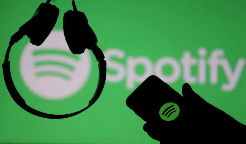 A smartphone and a headset are seen in front of a screen projection of Spotify logo, in this picture illustration