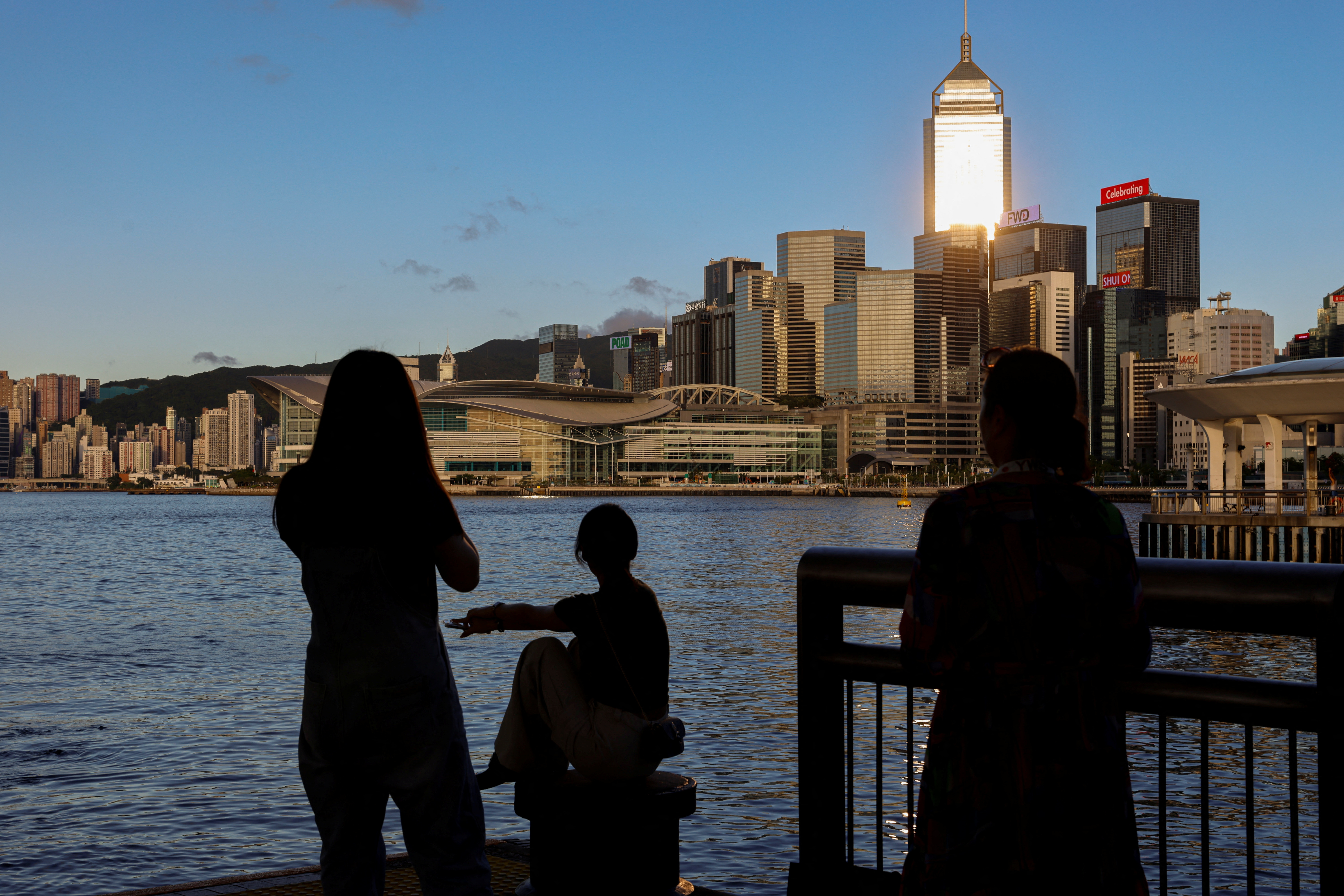 Tourists relax by the Victoria Harbour waterfront, with the iconic skyline providing a scenic backdrop, in Hong Kong