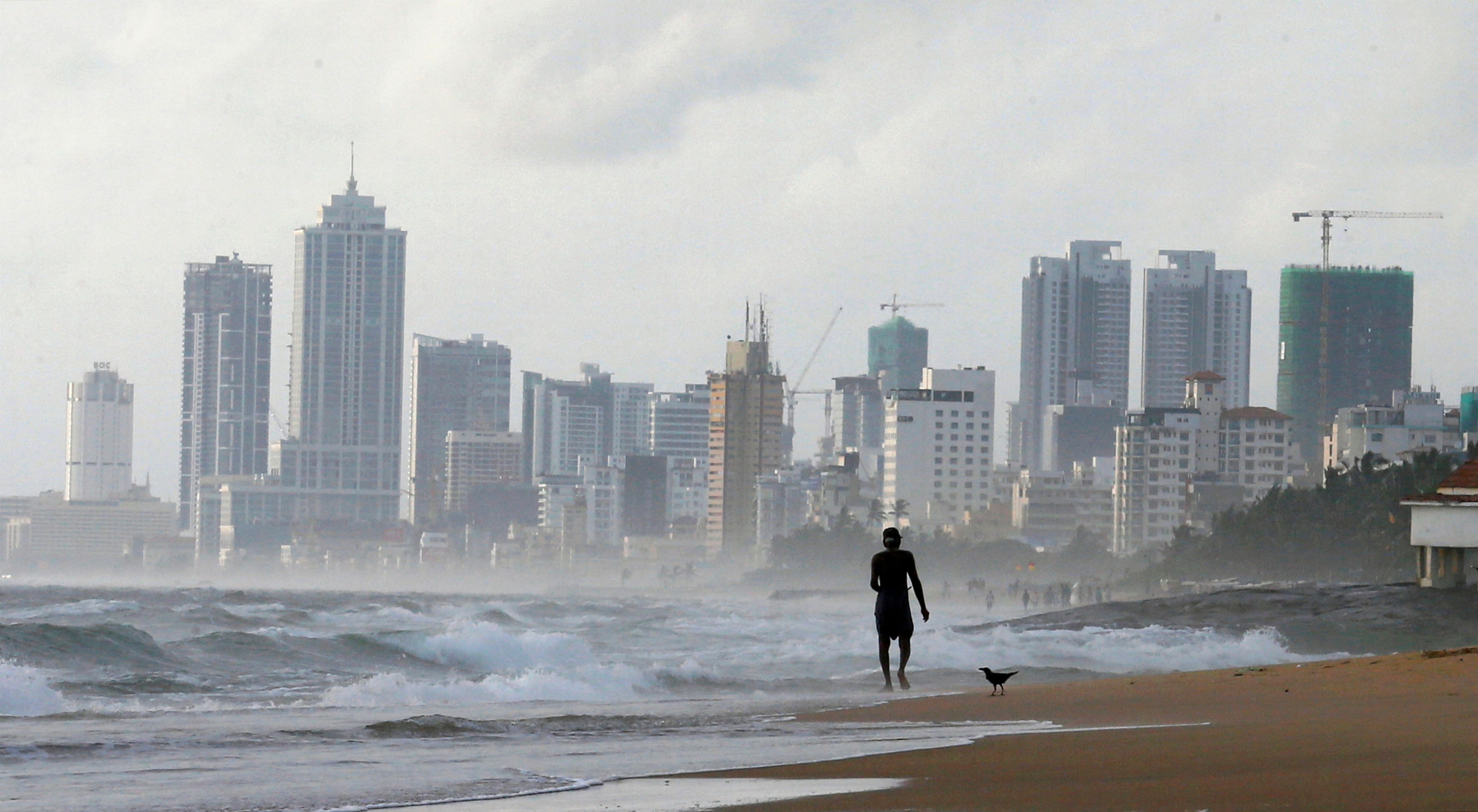 A man walks along a beach, against the backdrop of Colombo's Financial City
