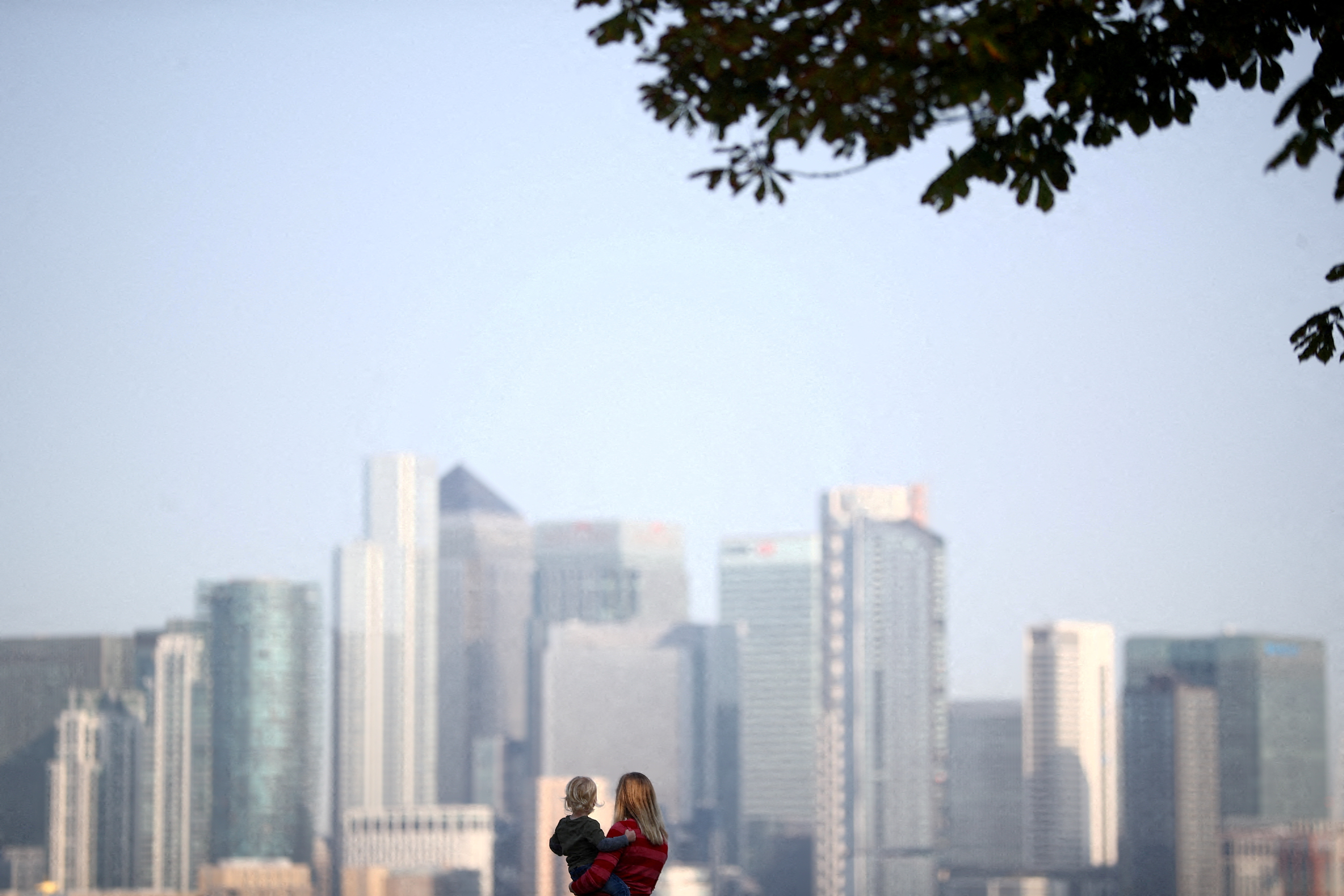 A woman holds a child in front of Canary Wharf skyline, in London