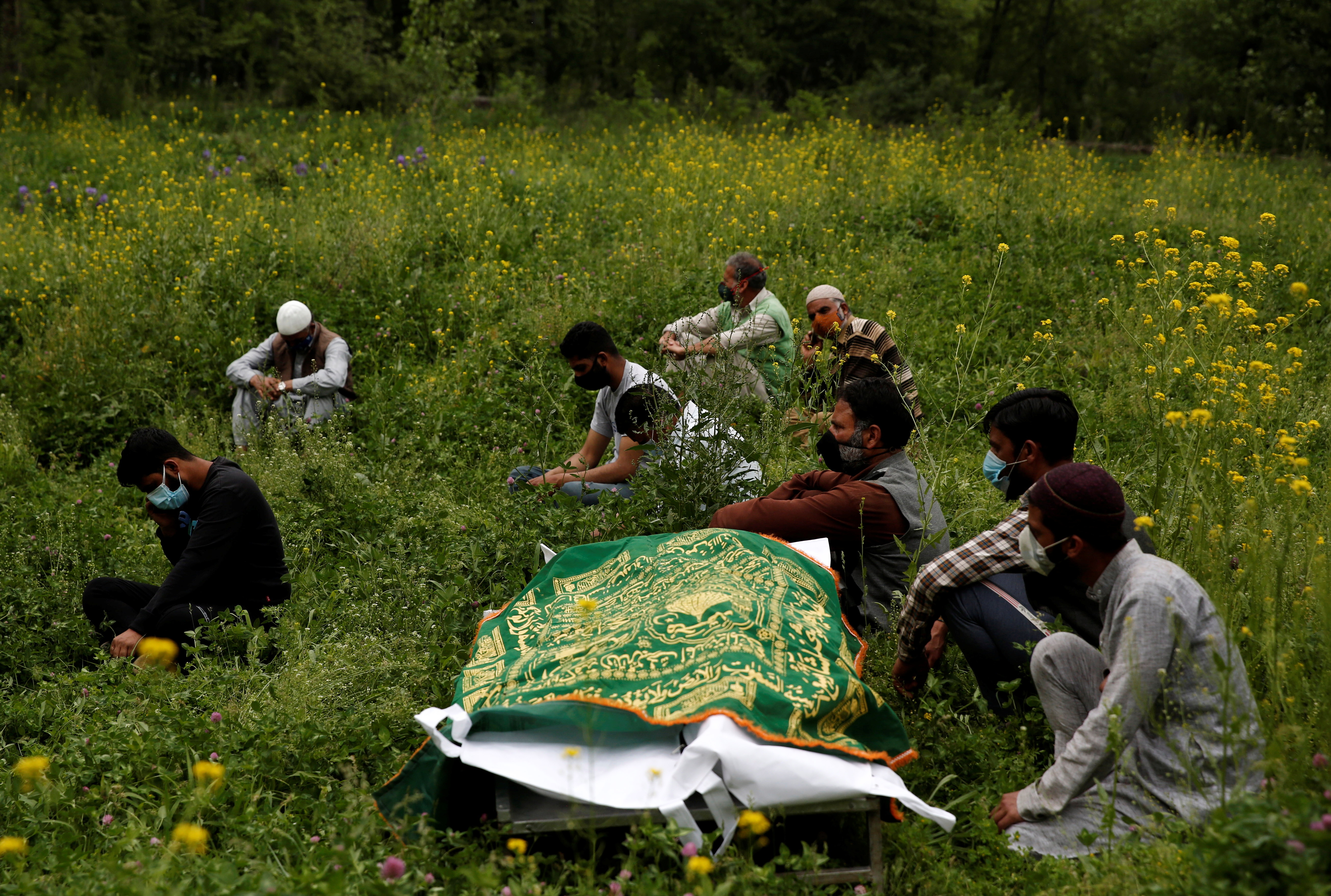 Relatives sit next to the body of a man, who died due to the coronavirus disease (COVID-19), as they wait for a grave to be prepared for his burial at a graveyard on the outskirts of Srinagar