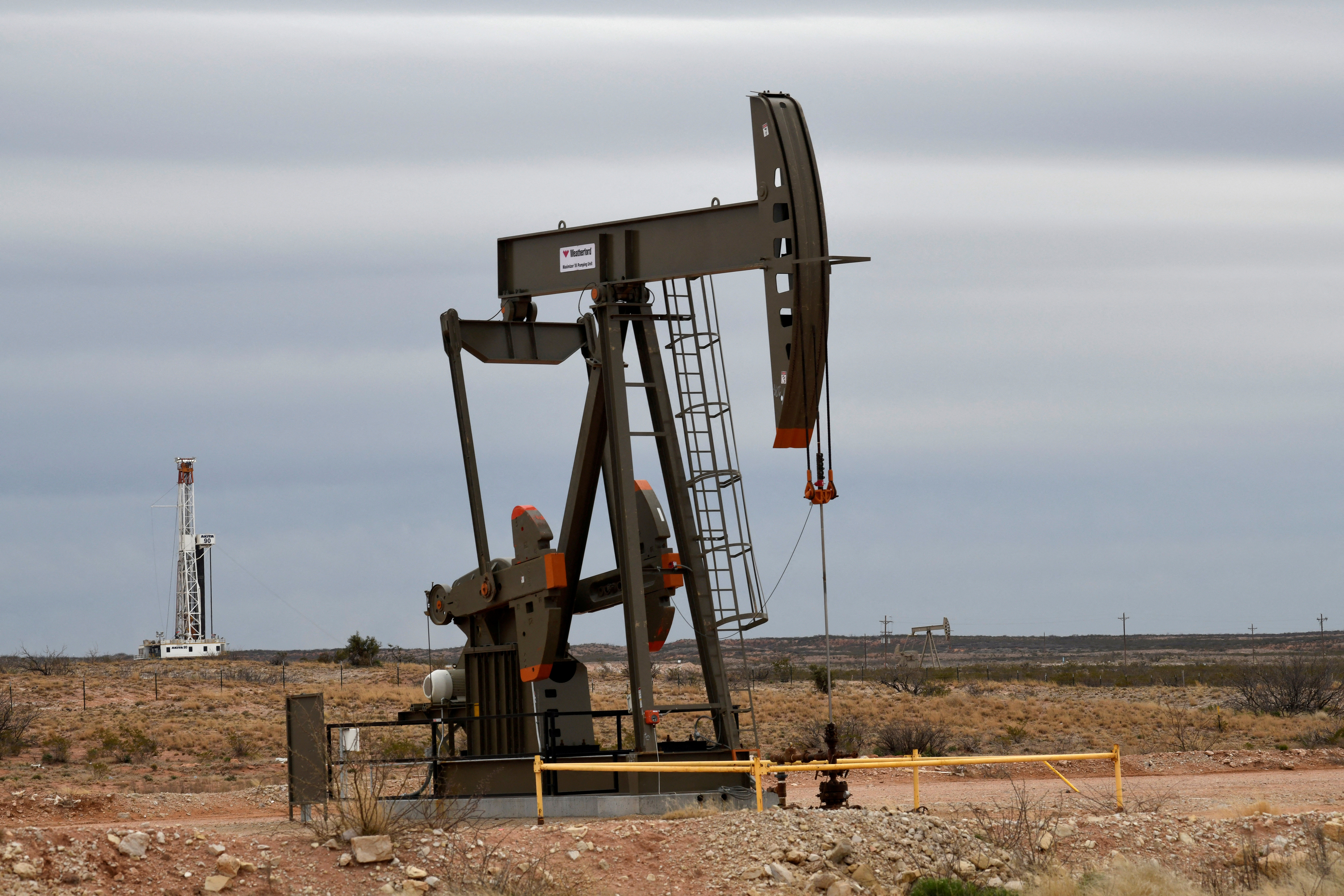 A pump jack operates in front of a drilling rig owned by Exxon near Carlsbad