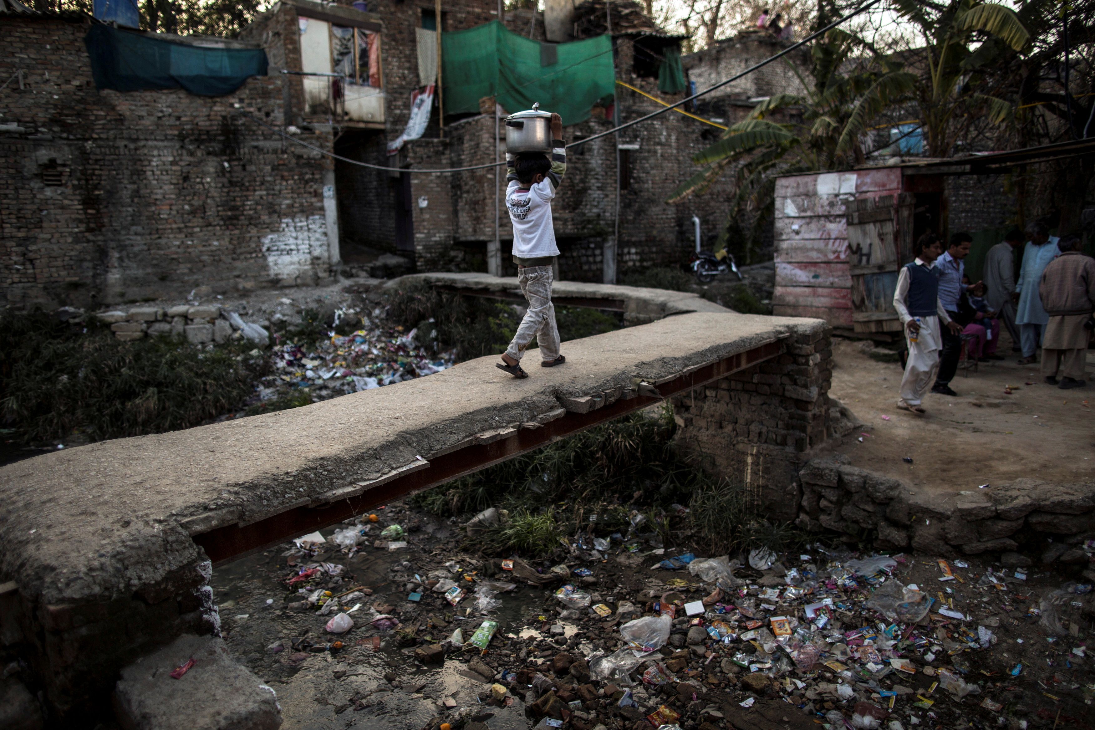 A boy carries a cooking pot on his head as he walks above an open sewer at a slum in Islamabad