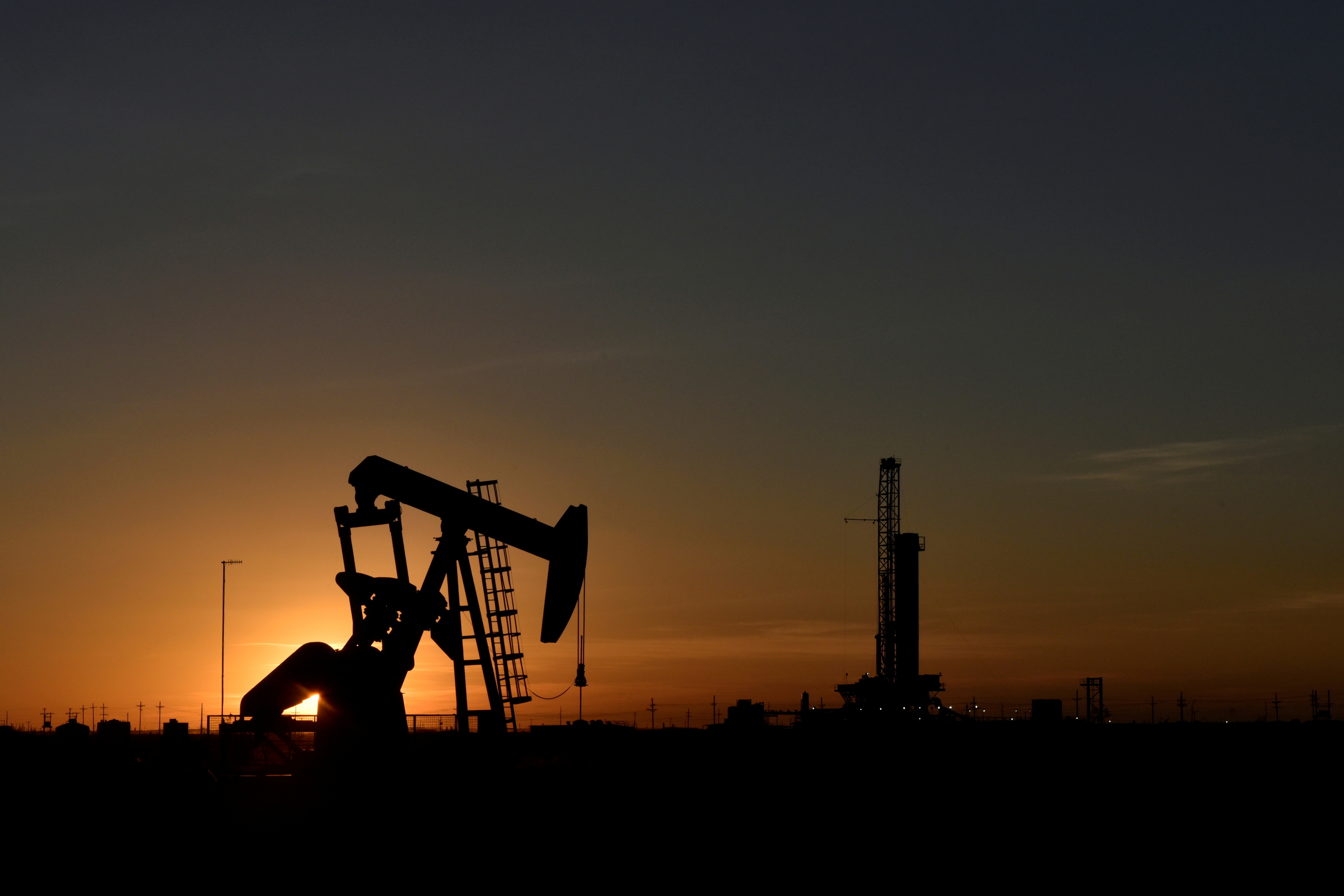 A pump jack operates in front of a drilling rig at sunset in an oil field in Midland