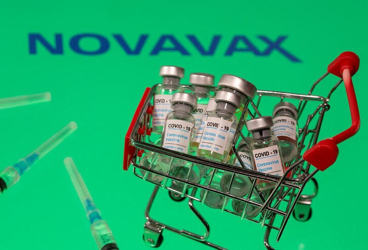 A small shopping basket filled with vials labeled "COVID-19 - Coronavirus Vaccine" and medical sryinges are placed on a Novavax logo