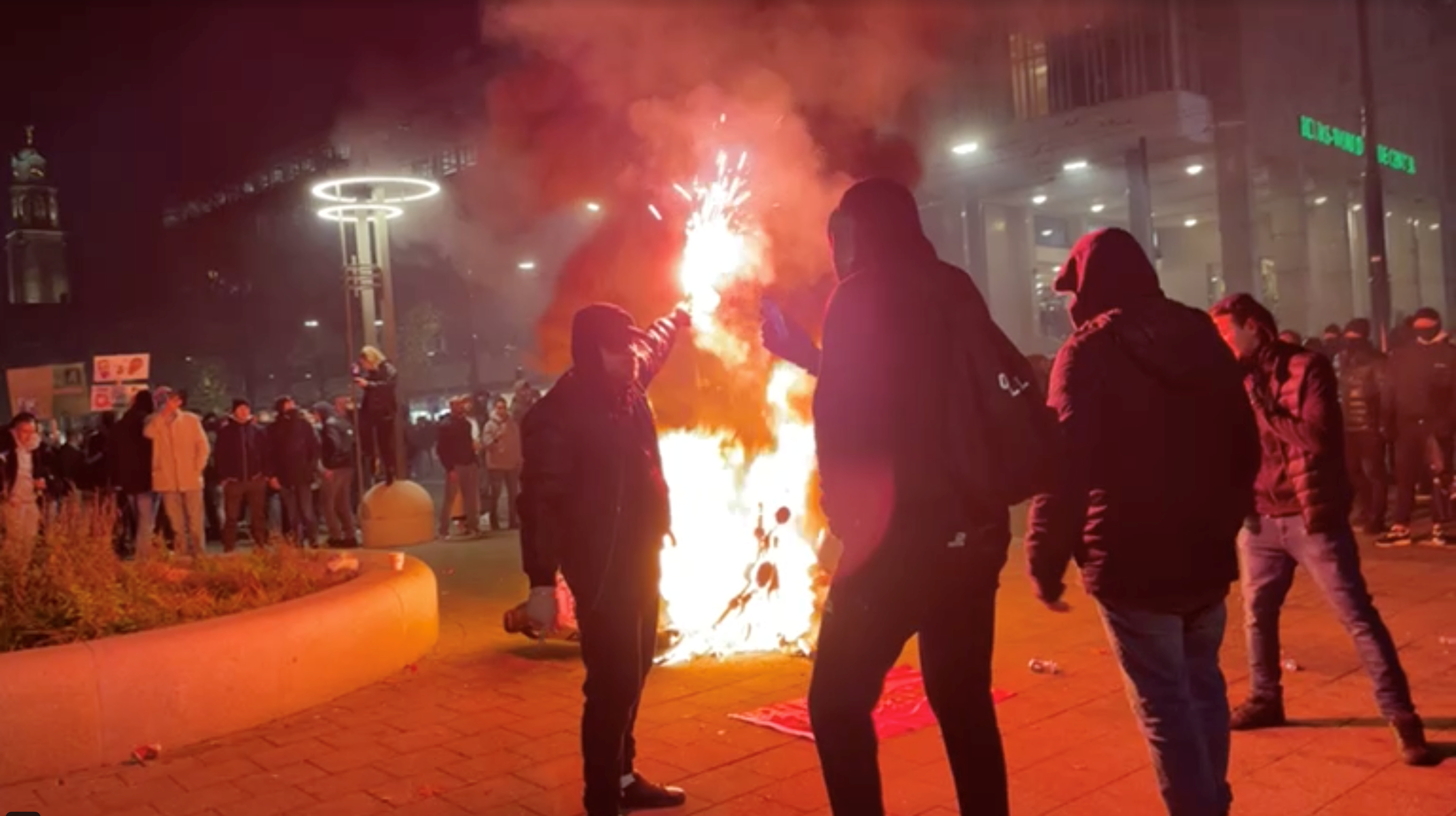 A man stands with a flare as protesters watch a motorcycle burning in Rotterdam