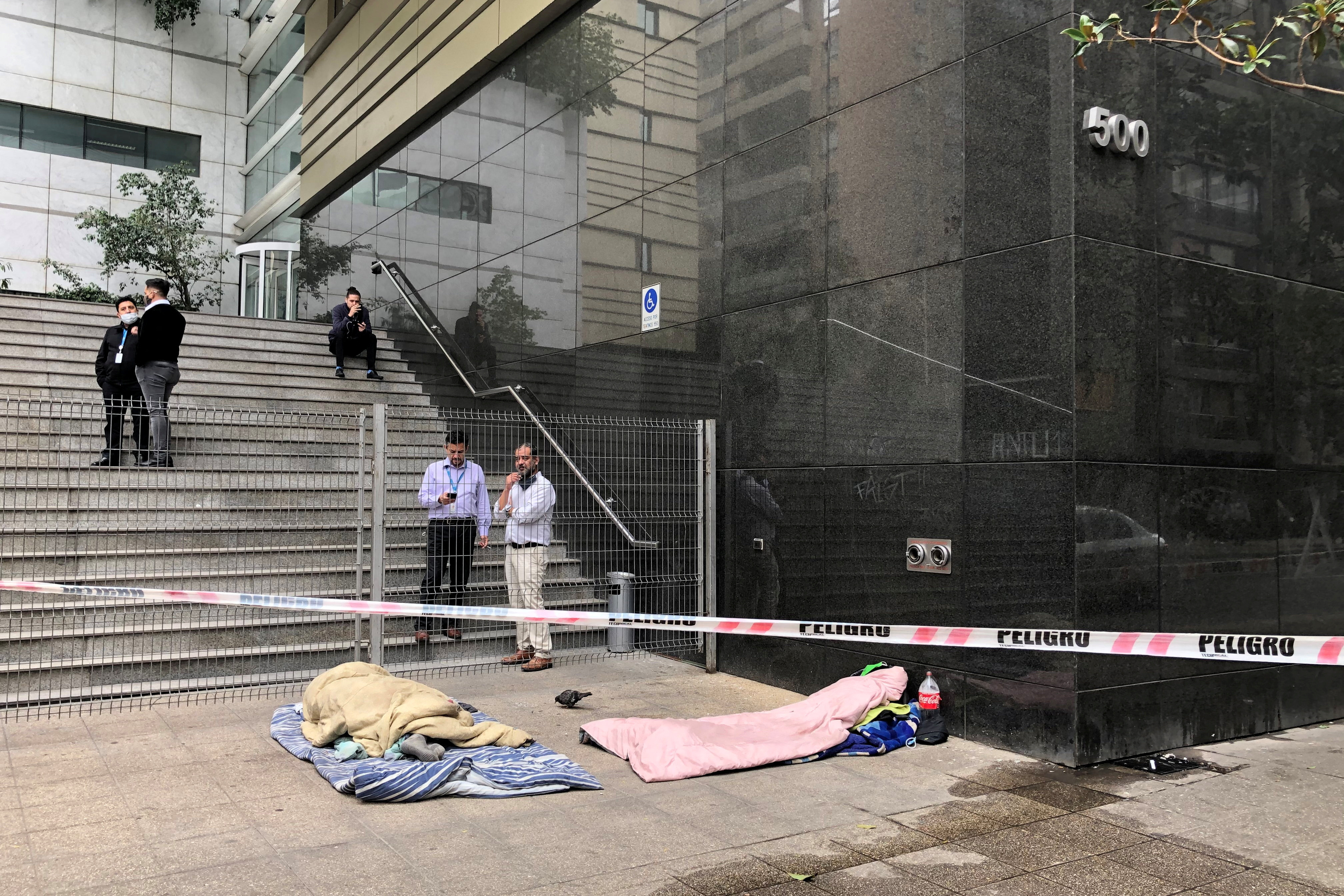 Workers take a break as homeless people sleep behind barricade tape outside an office building in downtown Santiago