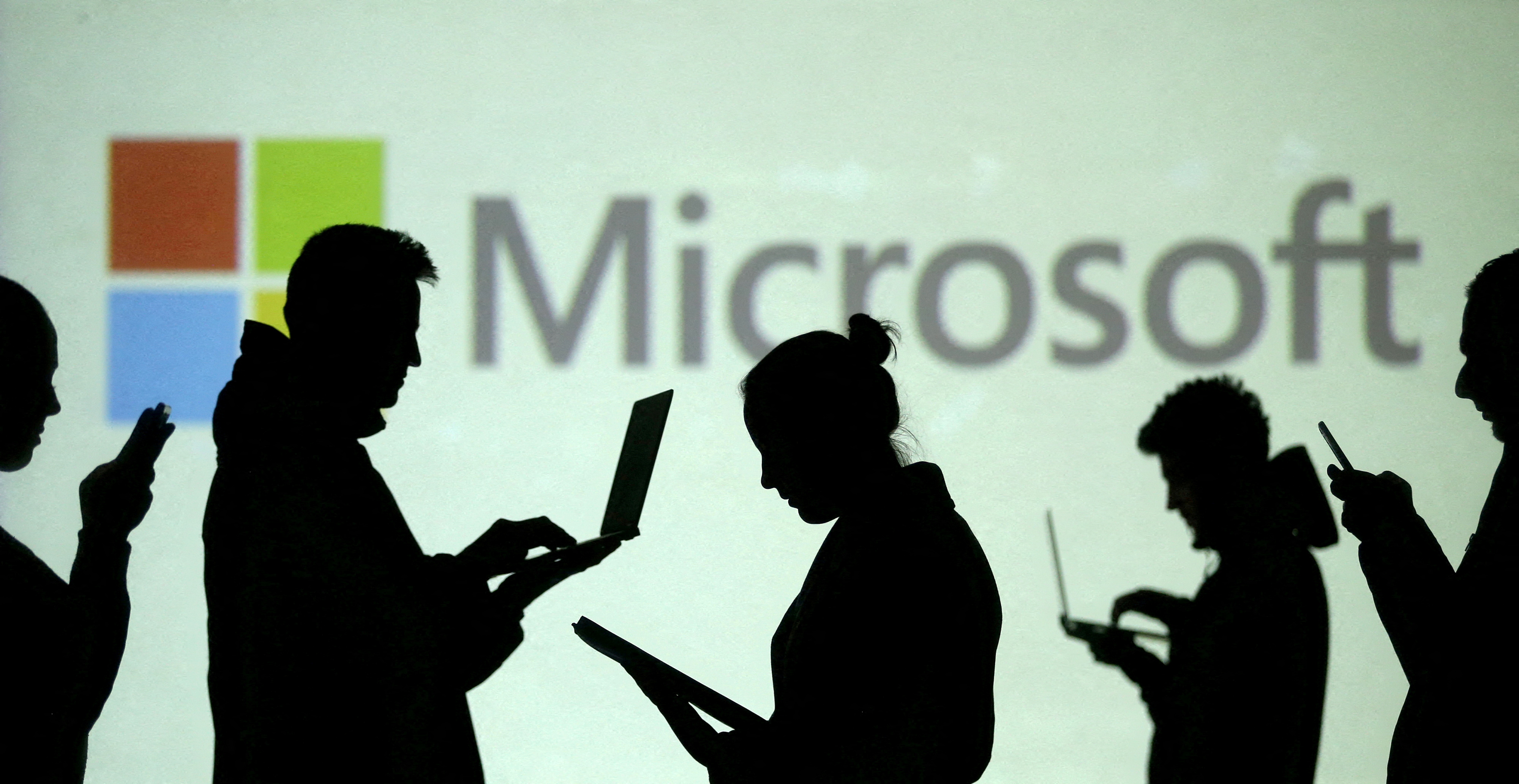 Silhouettes of laptop and mobile device users are seen next to a screen projection of Microsoft logo in this picture illustration