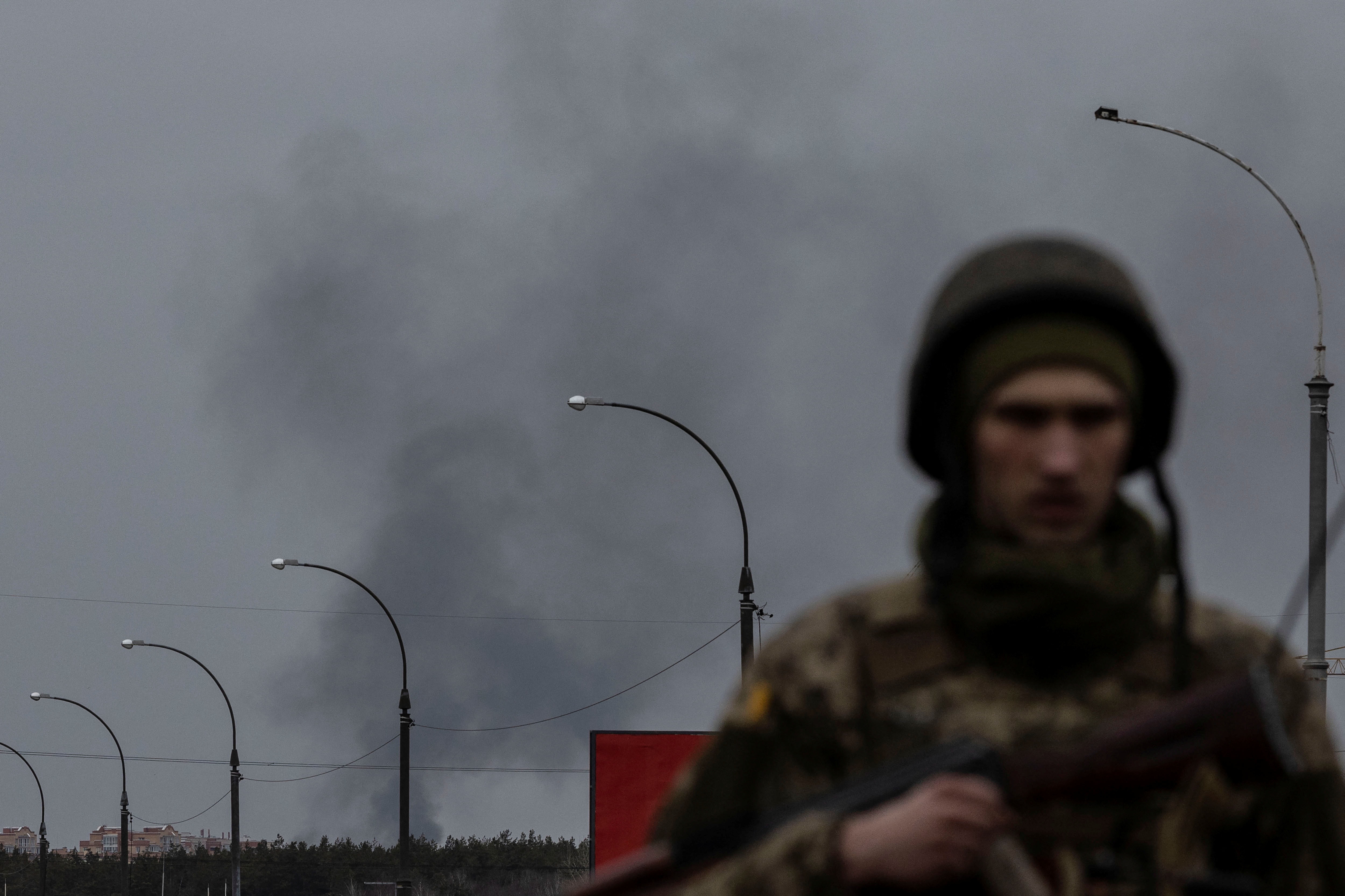 Smoke rises as a service member of the Ukrainian armed forces stands by the only escape route used by locals to evacuate from the town of Irpin, after days of heavy shelling, while Russian troops advance towards the capital, in Irpin, near Kyiv