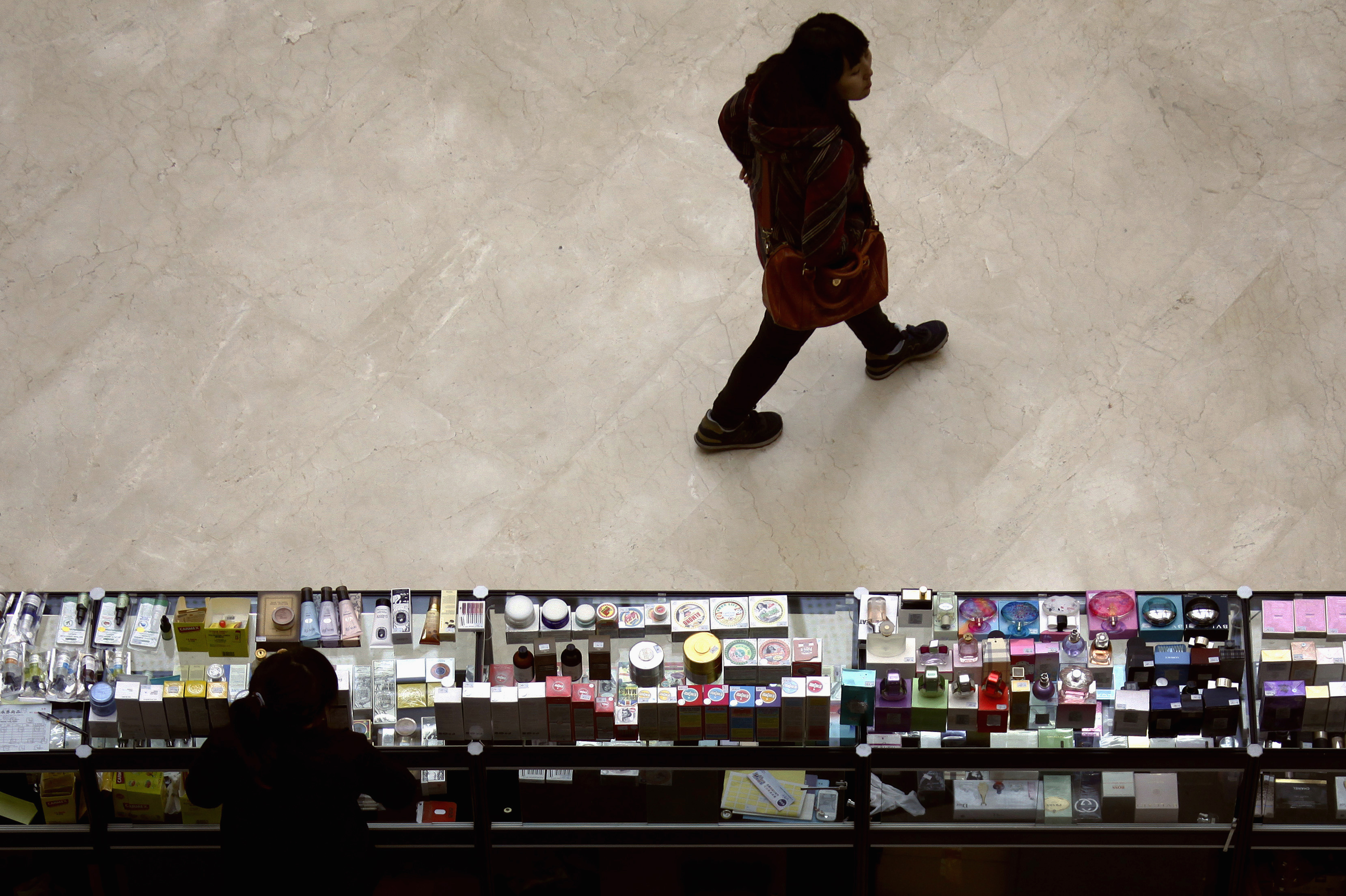 A shopper walks past a woman at a counter displaying cosmetics at a shopping mall in Beijing