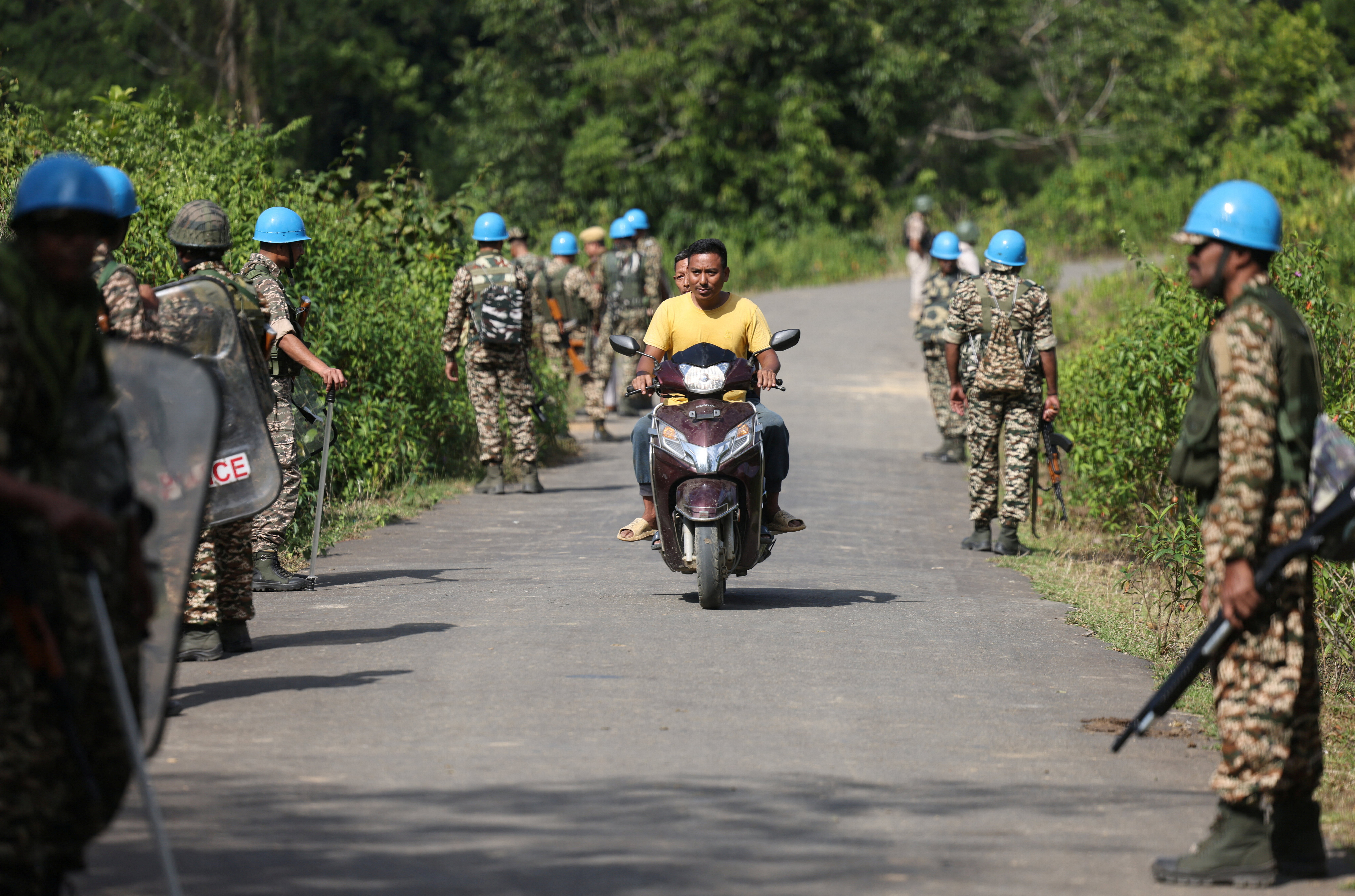 People ride a scooter past Central Reserve Police Force personnel standing guard before the funeral of Meiteis who were killed after ethnic violence broke out in Borobekra, Jiribam