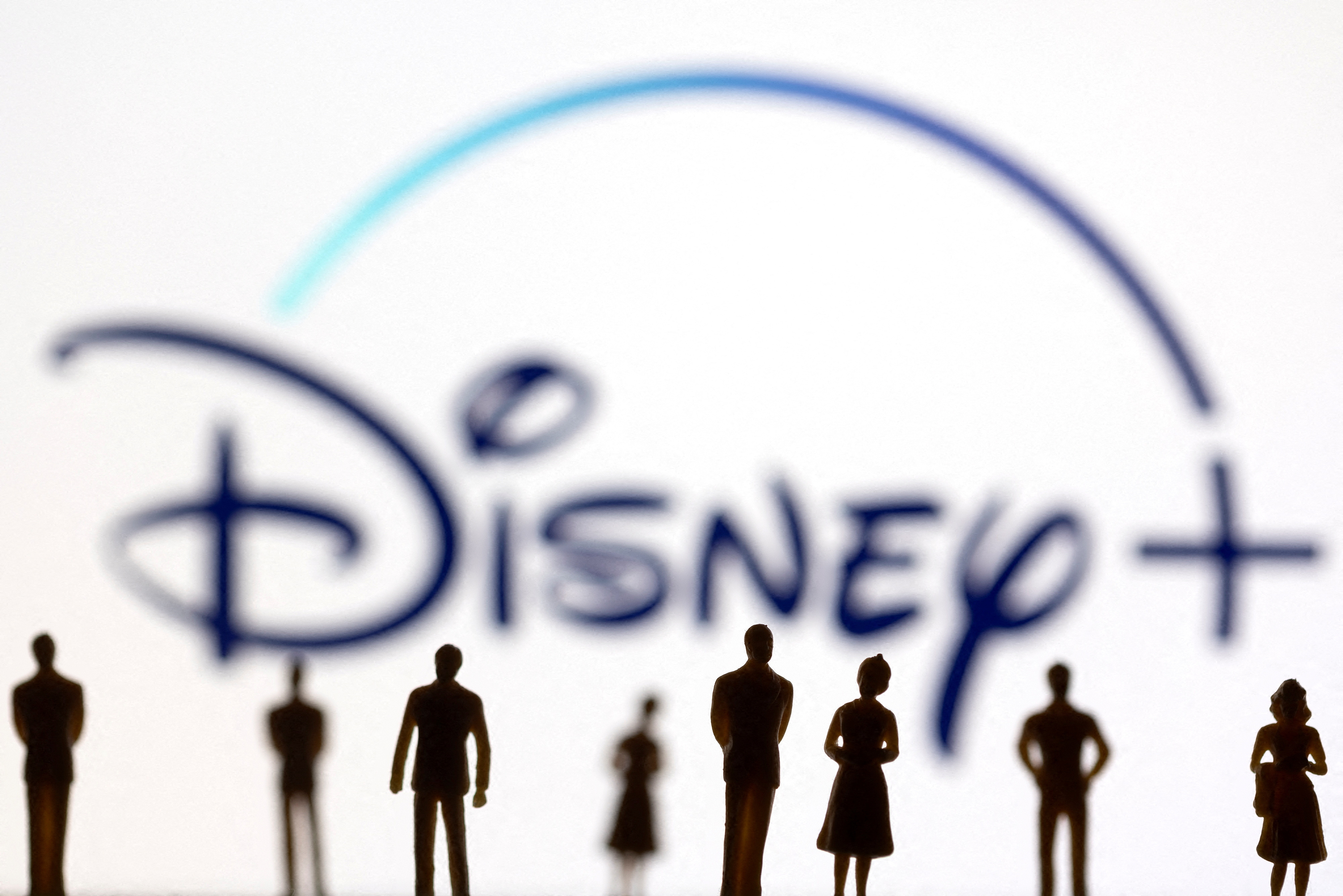 Toy figures of people are seen in front of the displayed Disney + logo, in this illustration