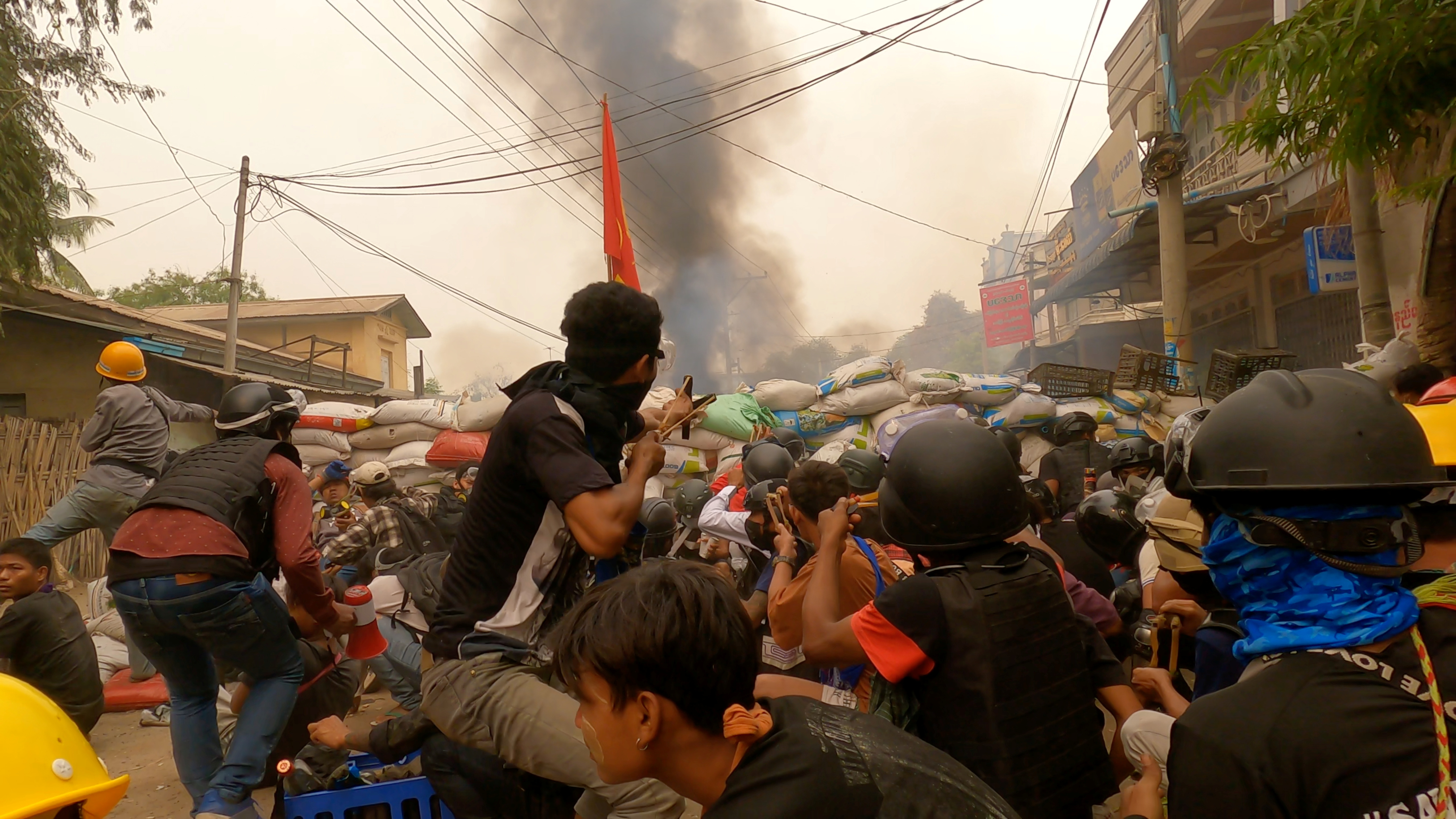 Protesters use slingshots while taking cover behind a barricade as smoke rises from burning debris during ongoing protests against the military coup, in Monywa, Sagaing region, Myanmar March 29, 2021 in this still image from video obtained by REUTERS 