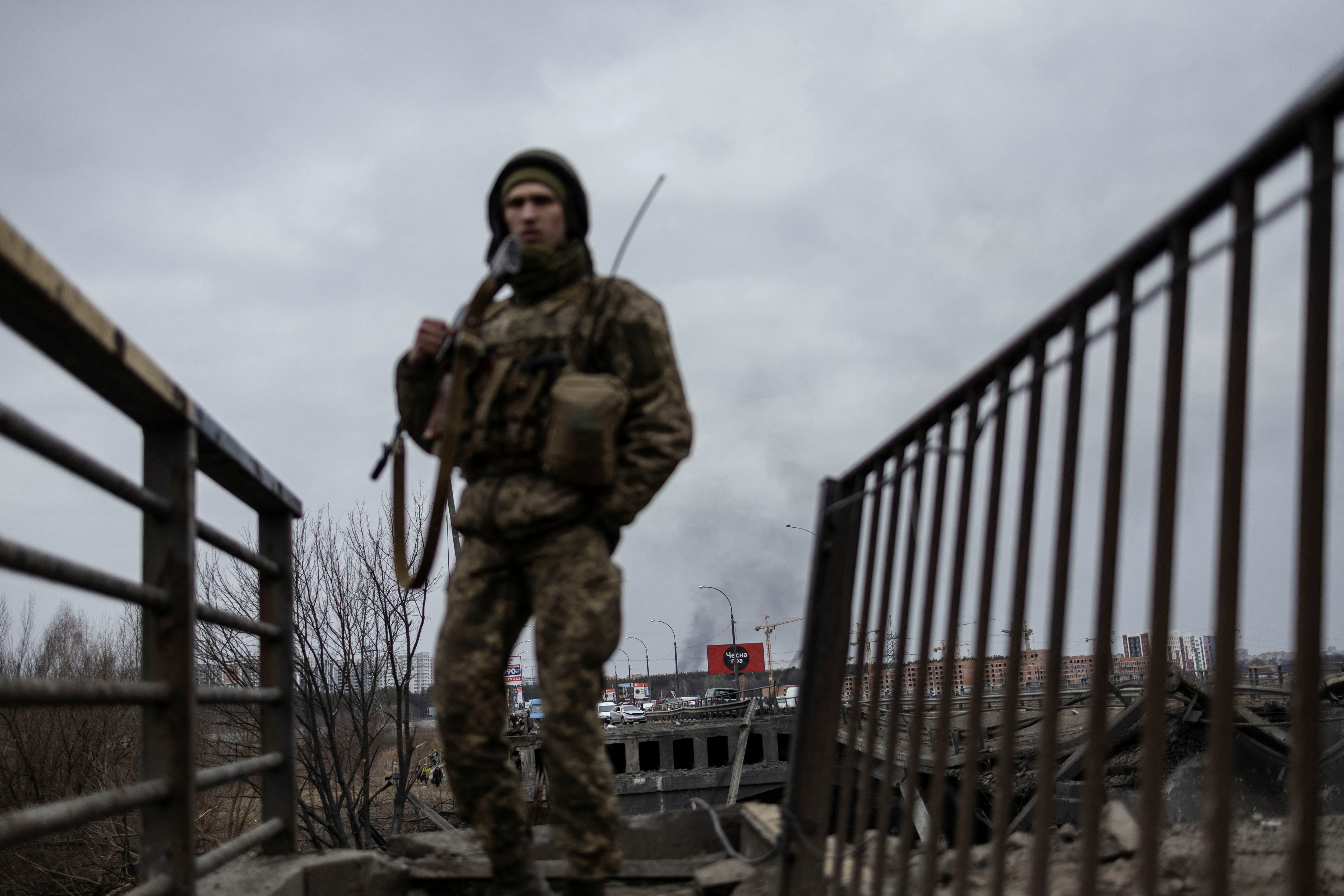 Smoke rises as a service member of the Ukrainian armed forces stands by the only escape route used by locals to evacuate from the town of Irpin, after days of heavy shelling, while Russian troops advance towards the capital, in Irpin, near Kyiv