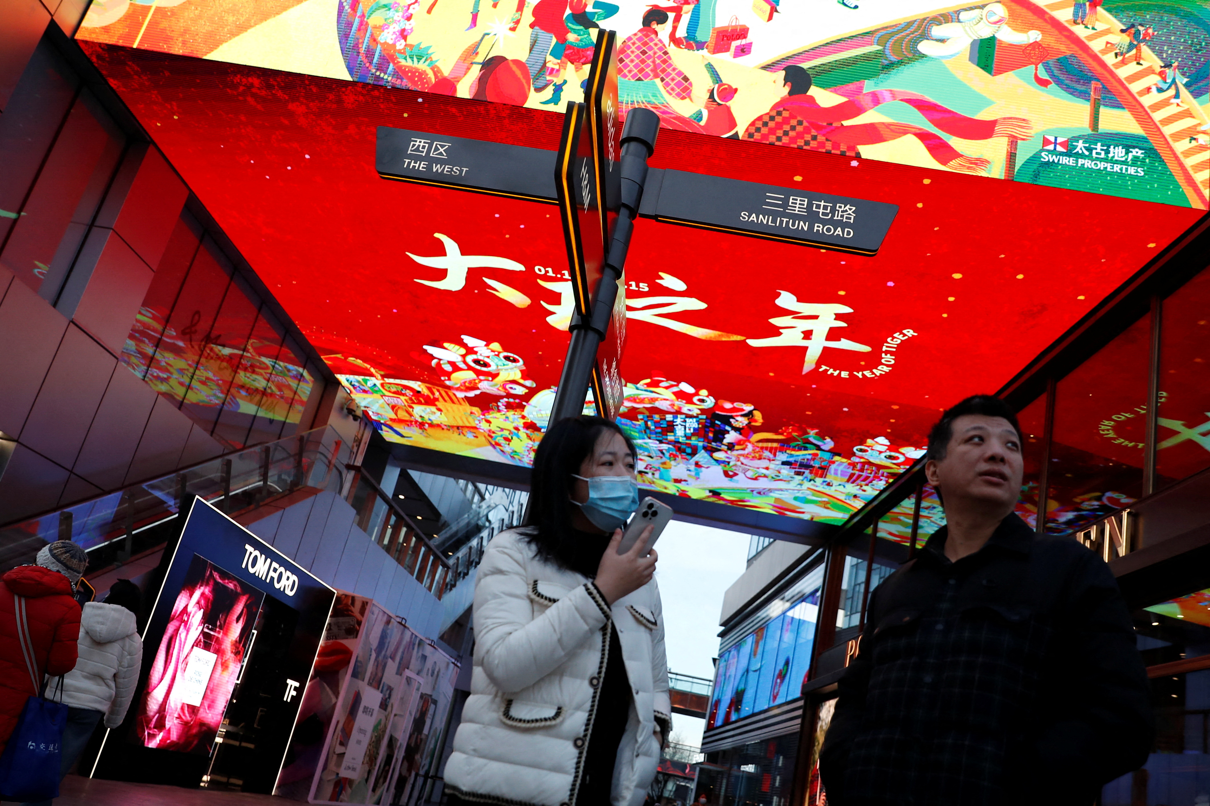 People stand under a giant screen showing sales promotion during the Chinese Lunar New Year holiday, ahead of the holiday at a shopping area in Beijing