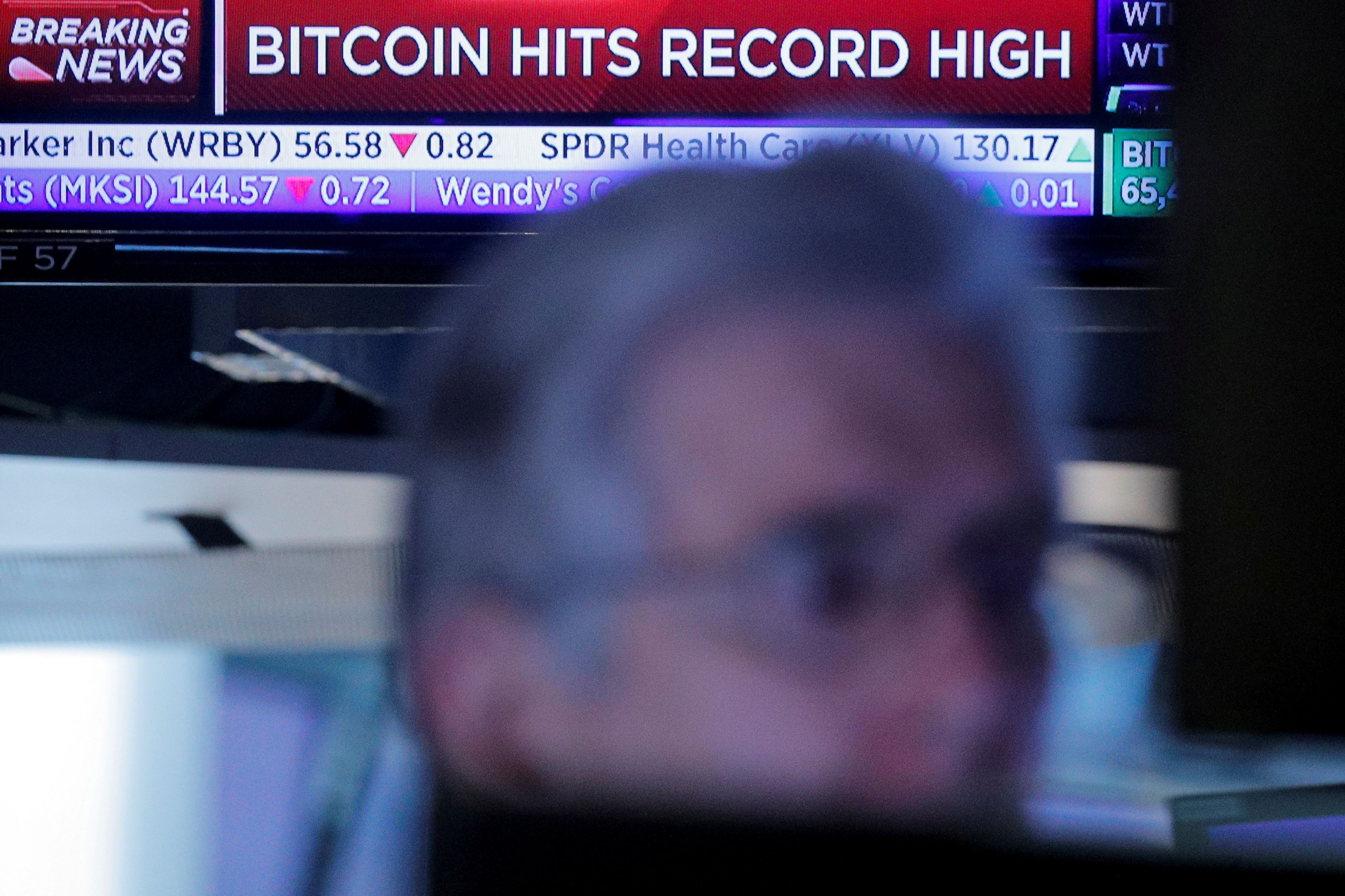 A screen displays a headline banner that reads; Bitcoin Hits Record High, as a trader works on the floor of the NYSE in New York
