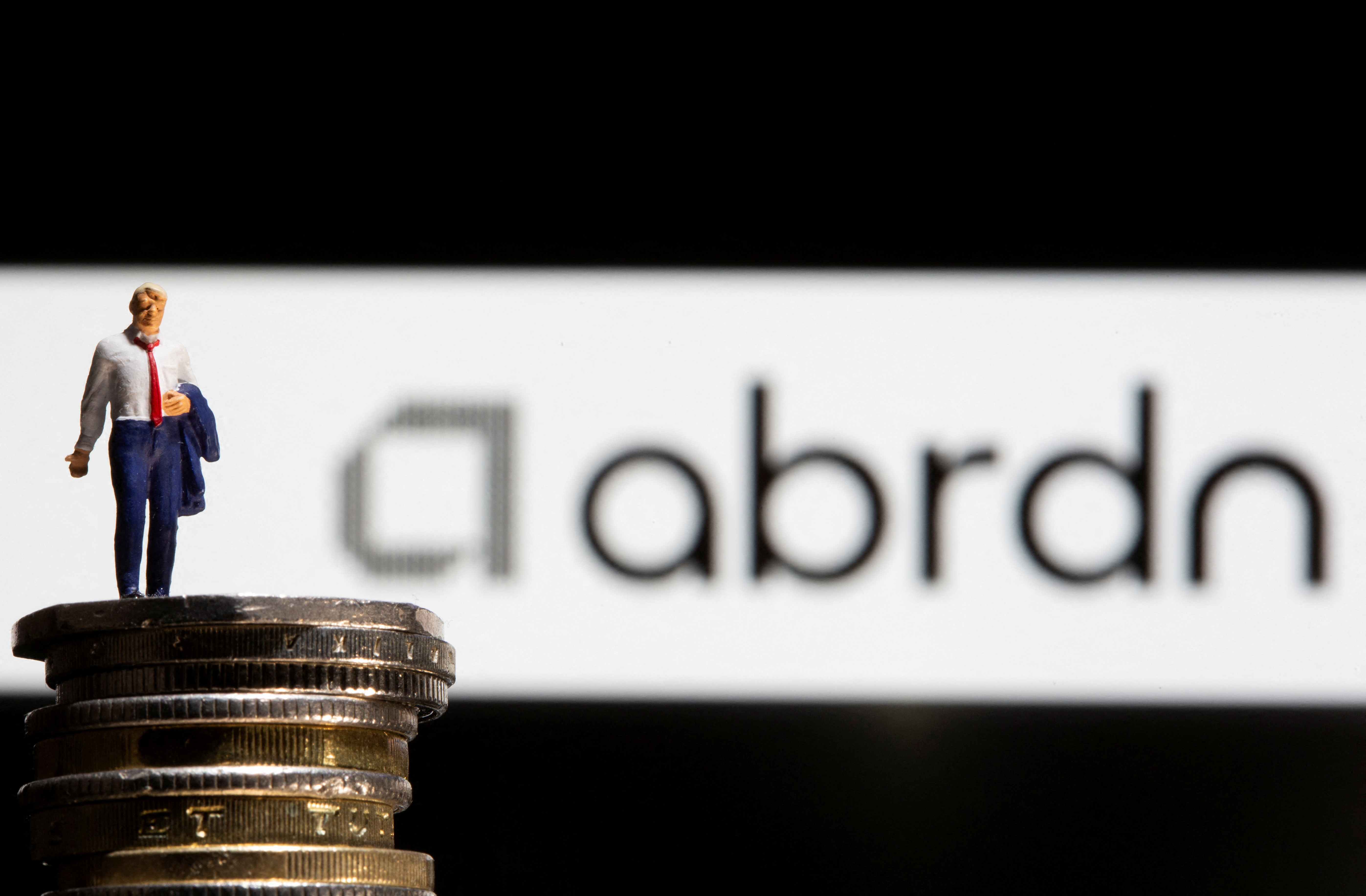 Businessman toy figure is placed on U.K. Pound coins in front of displayed Abrdn logo in this illustration taken