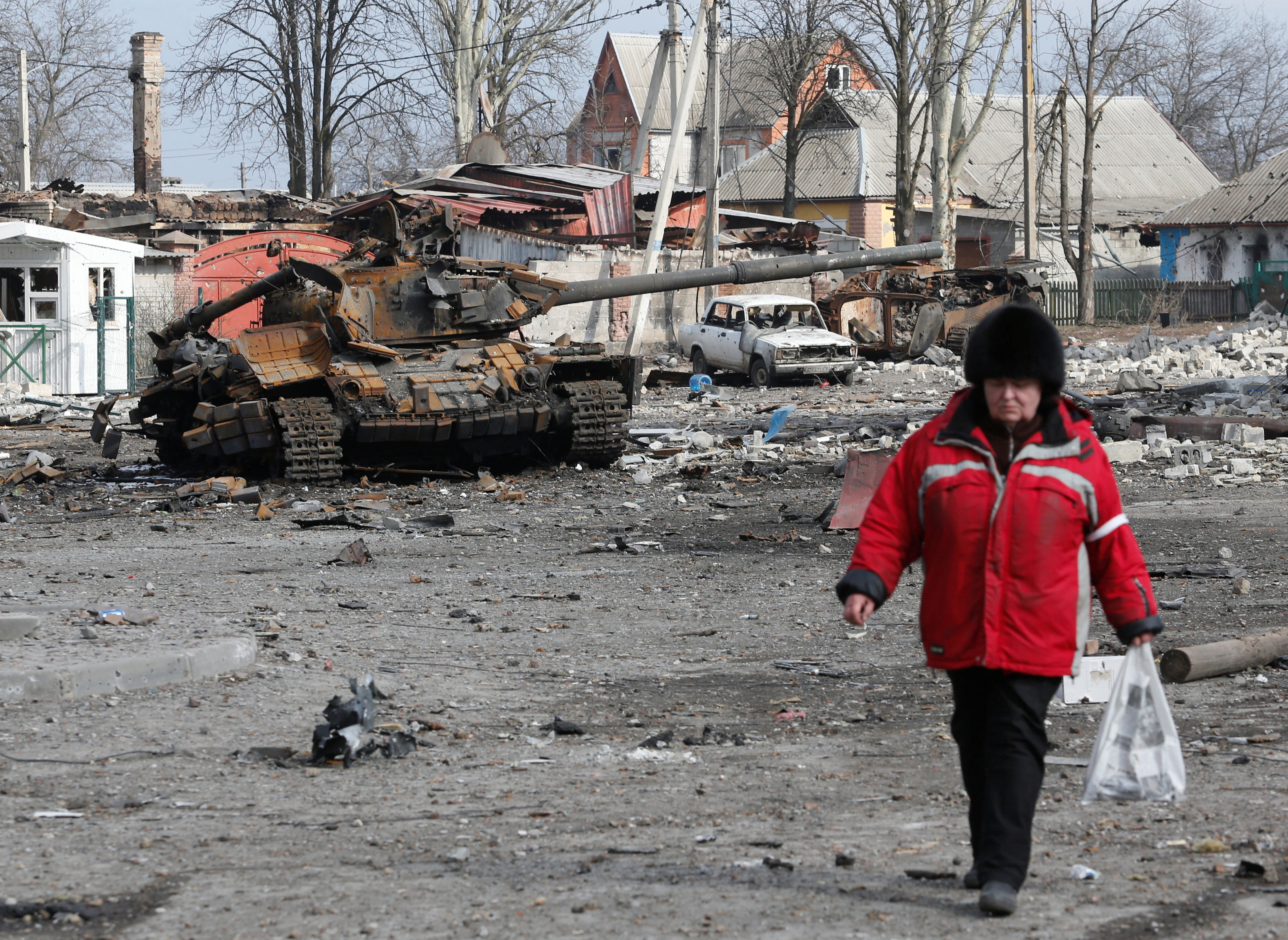 A woman walks past a destroyed tank in a damaged street in Volnovakha