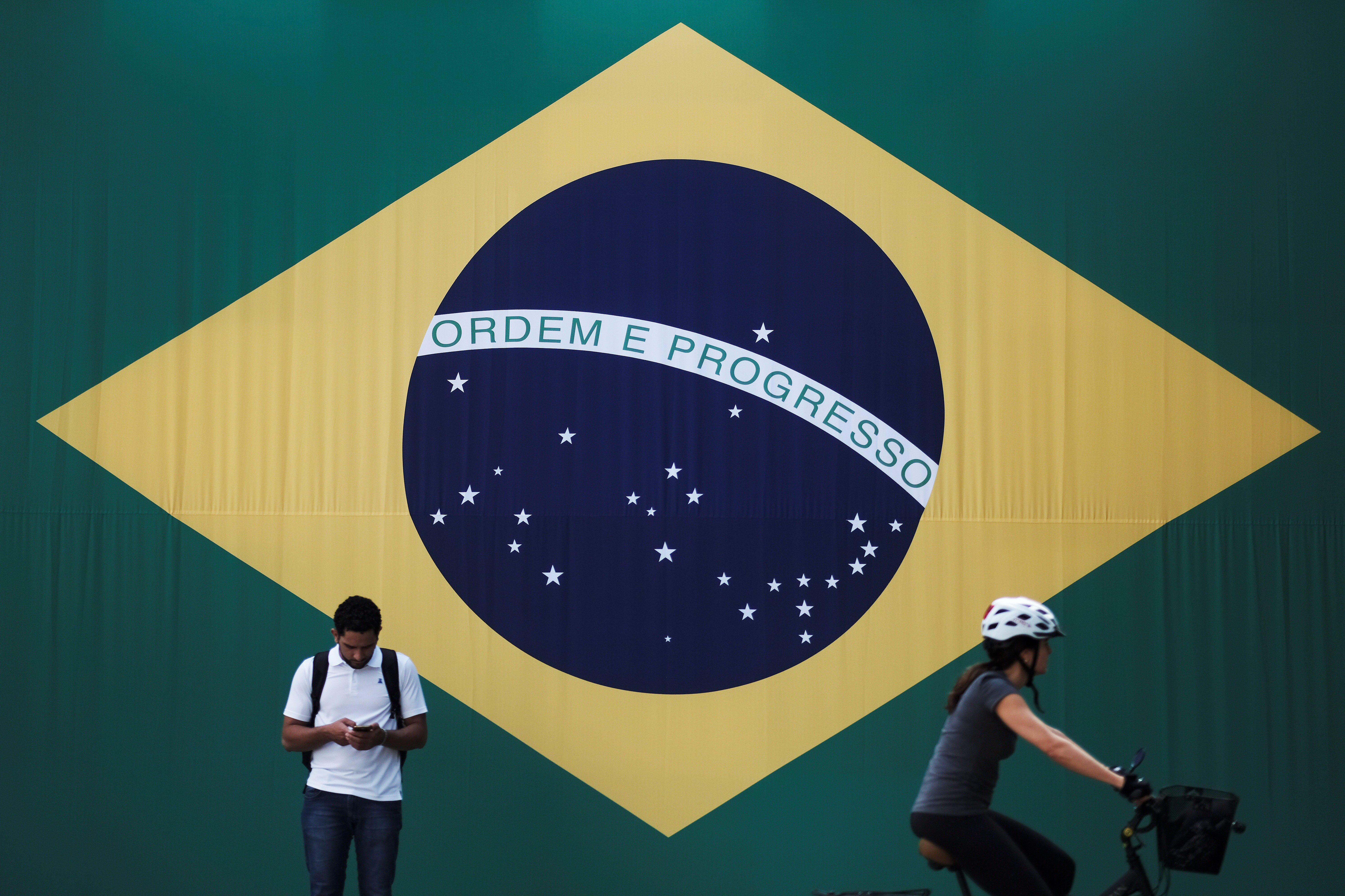 A man checks his mobile phone as a woman riding a bike passes next to a big Brazilian flag in Sao Paulo
