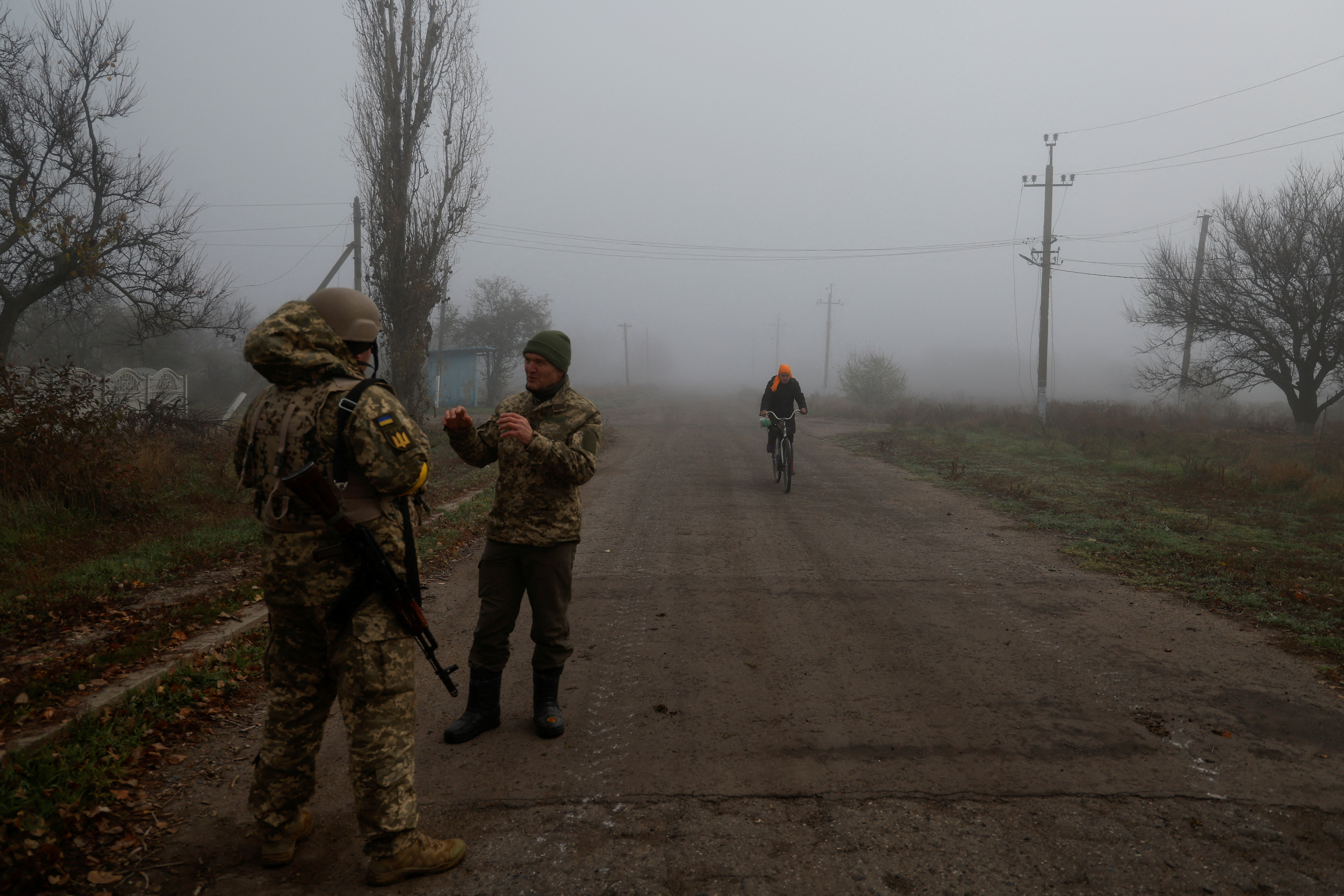 Ukrainian serviceman speaks with a chaplain while a local resident rides a bicycle along a street in a village near Shihurivka