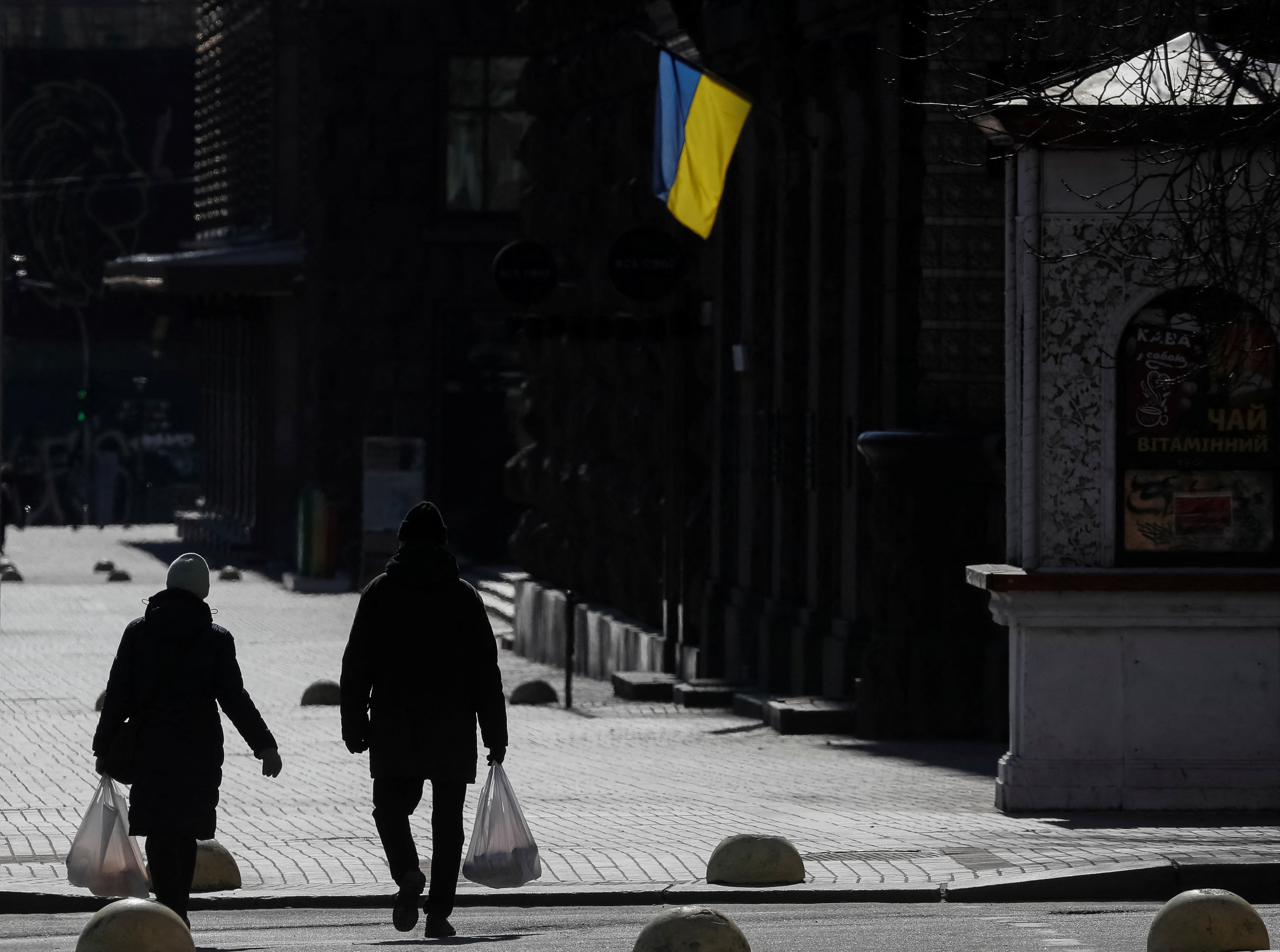 Locals residents walk on an empty street carrying bags with food, as an Ukrainian national flag is seen in central Kyiv