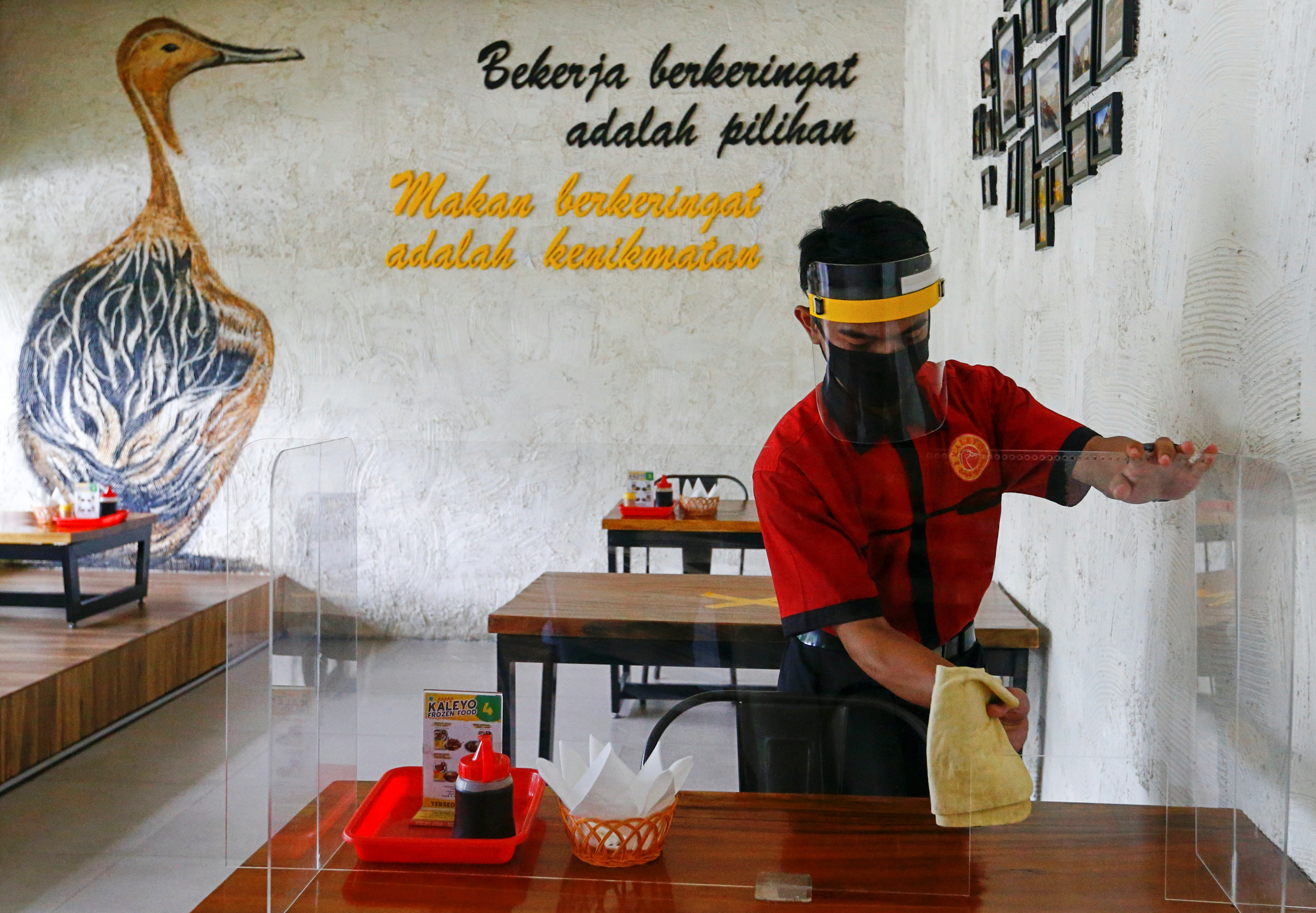 A worker wearing a face shield and a mask wipes a protective screen on a table at a restaurant, as the Indonesian capital kicks off a two-week 