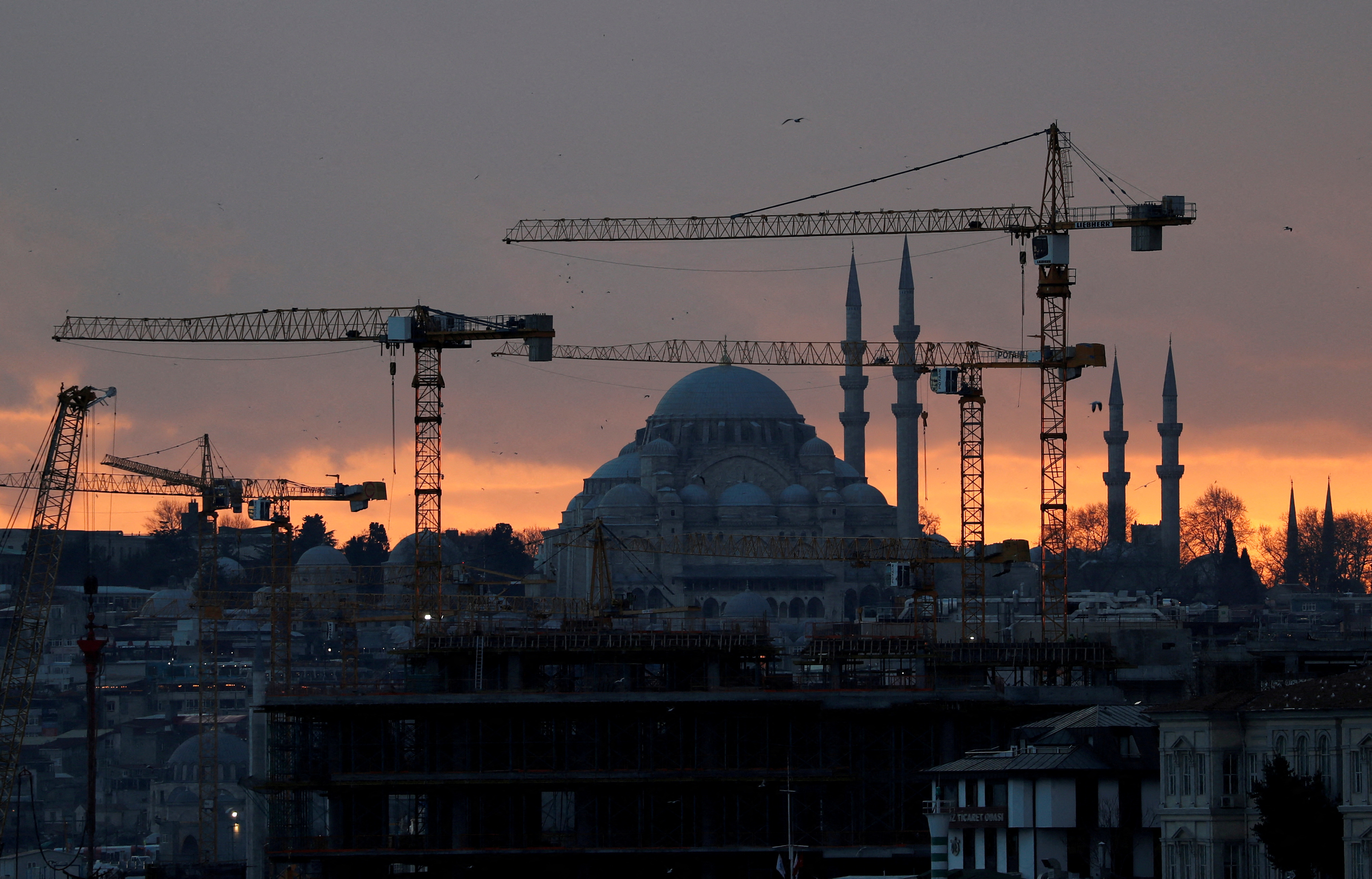 The sun sets behind cranes of Galata Port construction site and a mosque in Istanbul