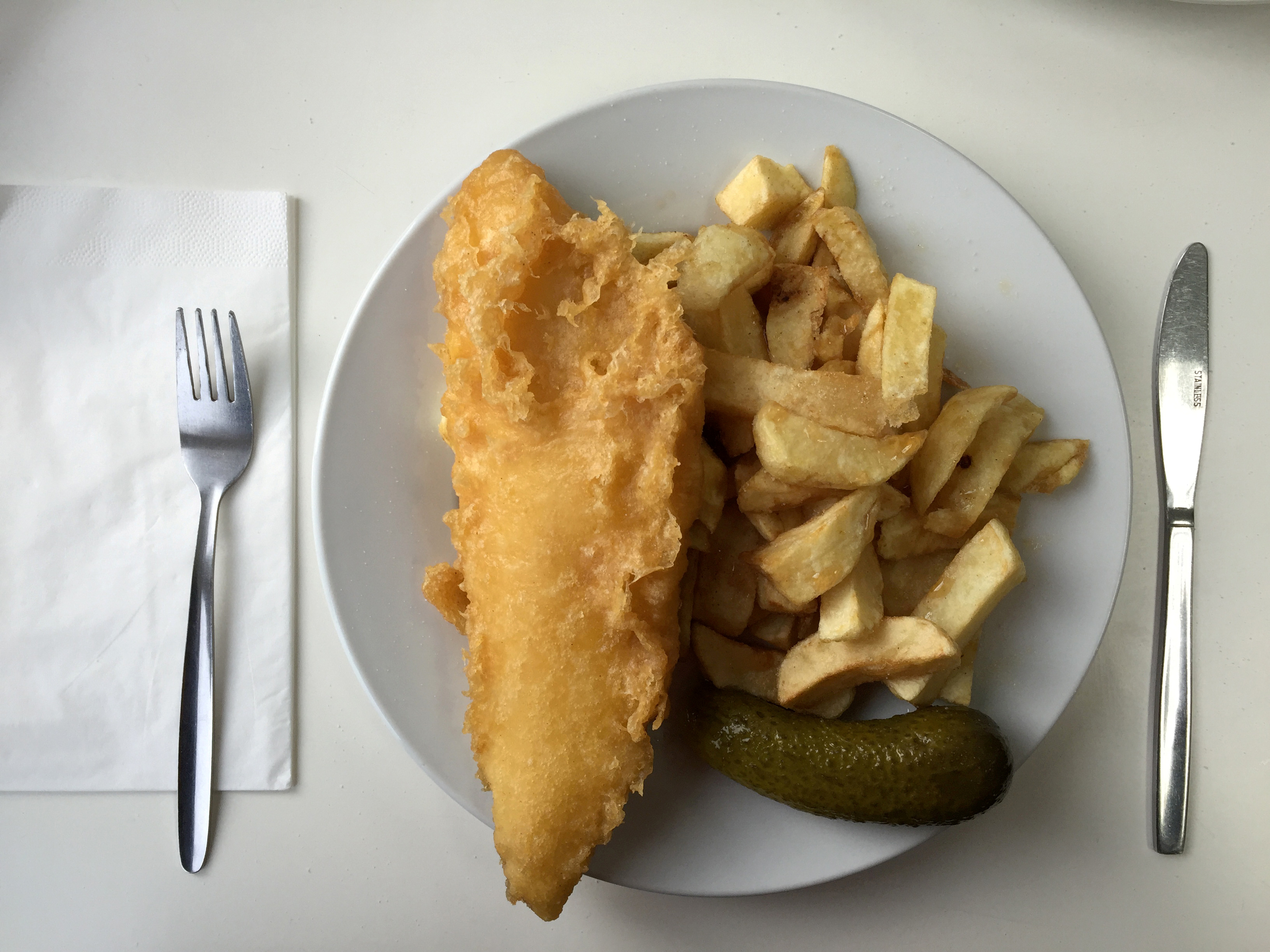 A plate of traditional fish and chips with a gherkin on a table in a seafront fish and chip shop in Brighton