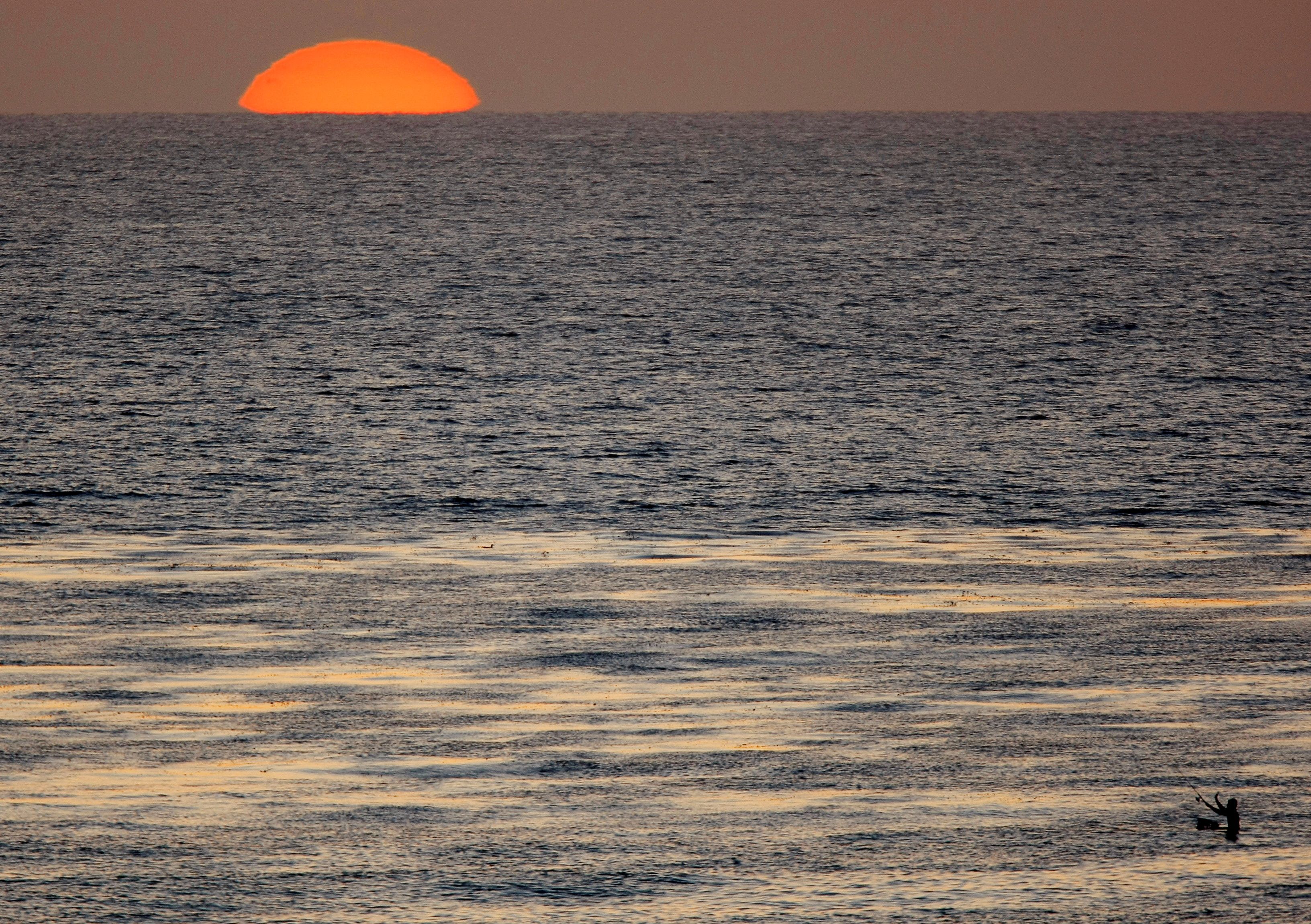 The sun sets as a fisherman casts his line in the Pacific Ocean while sitting on a surfboard off the coast of Cardiff