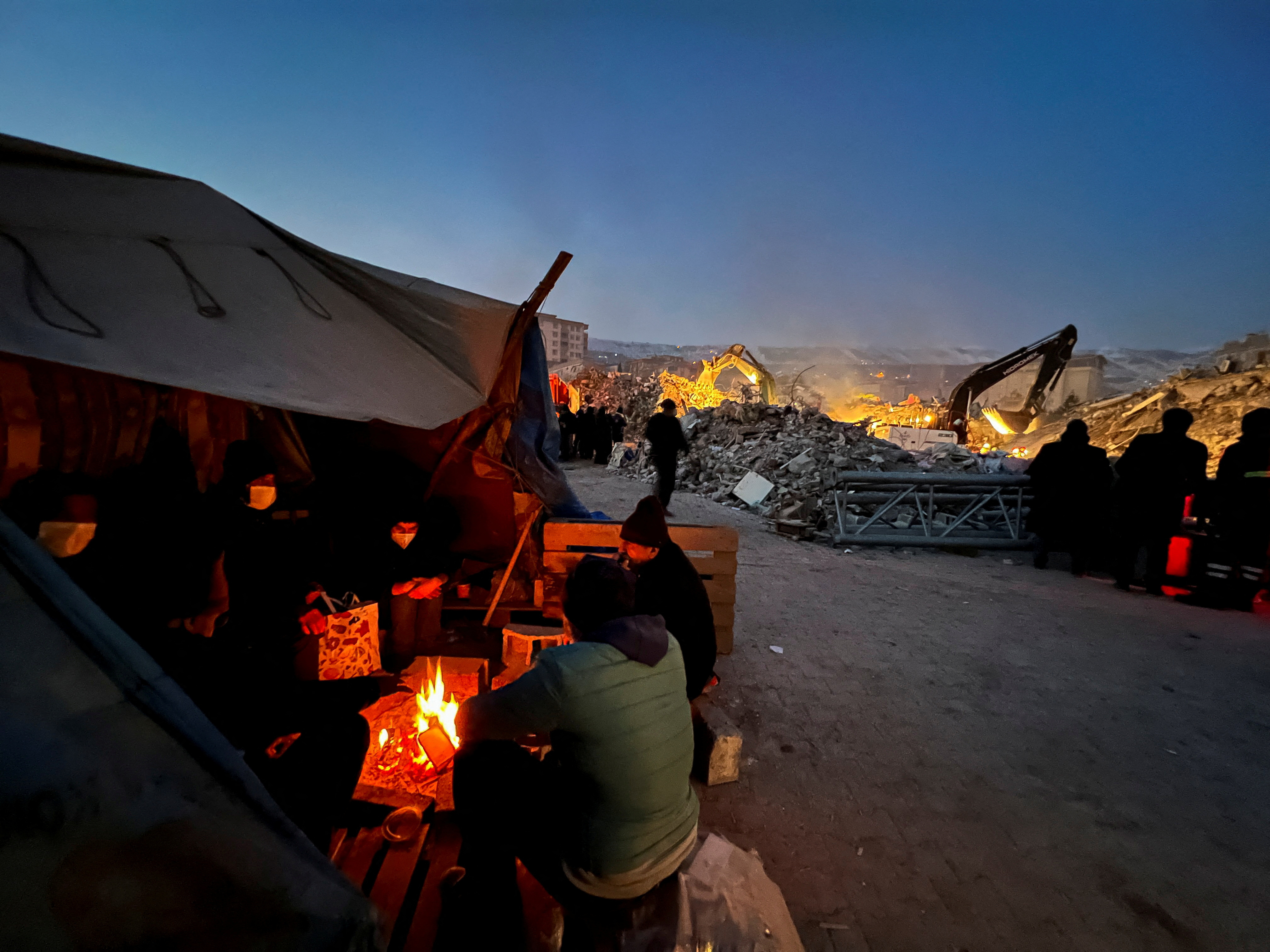 People sit around a fire near the site of a collapsed building in the aftermath of an earthquake, in Kahramanmaras