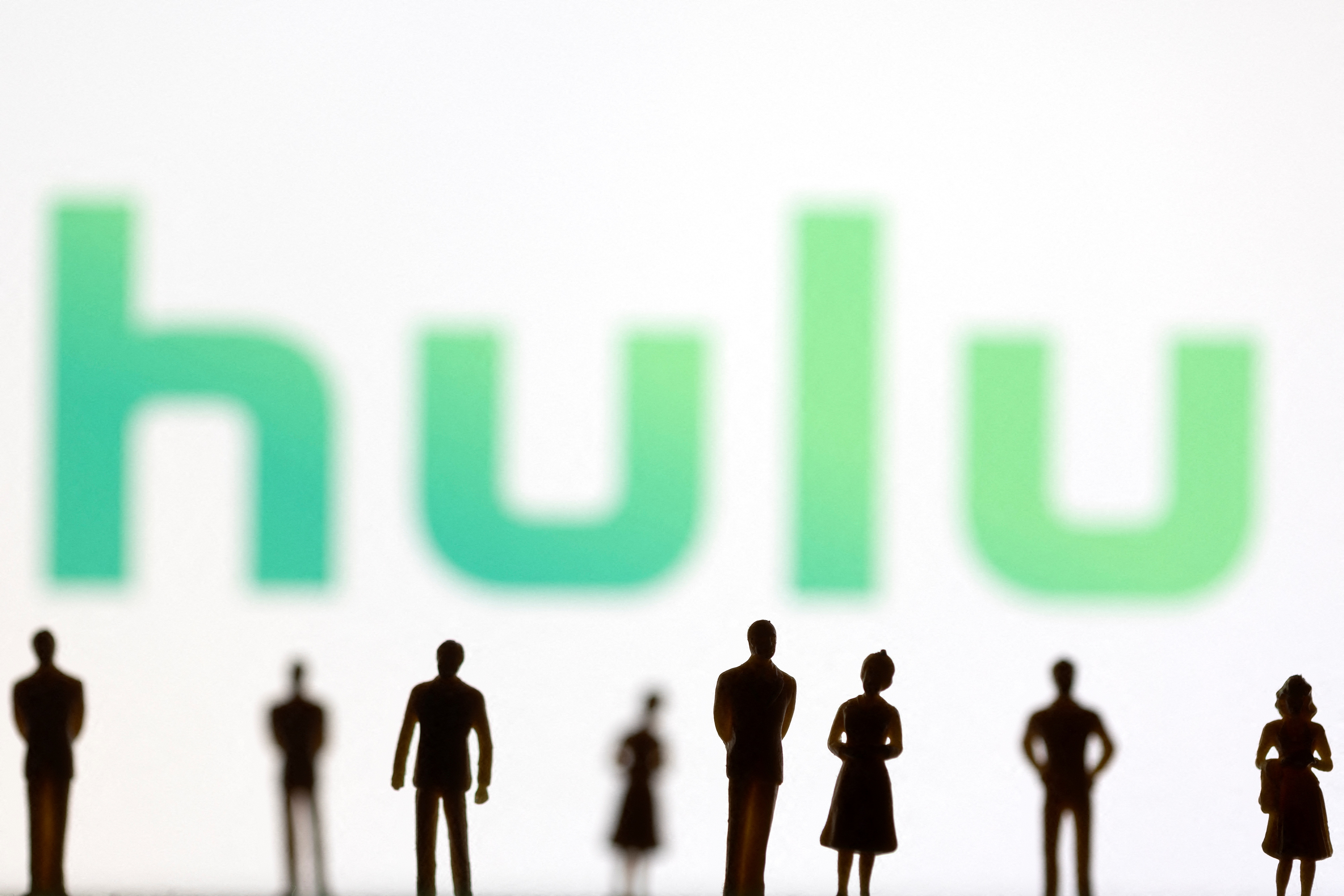 Toy figures of people are seen in front of the displayed Hulu logo, in this illustration