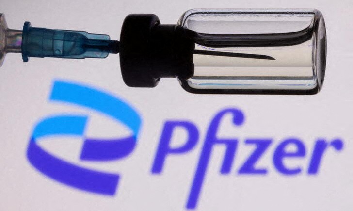 A vial and a syringe are seen in front of a displayed Pfizer logo