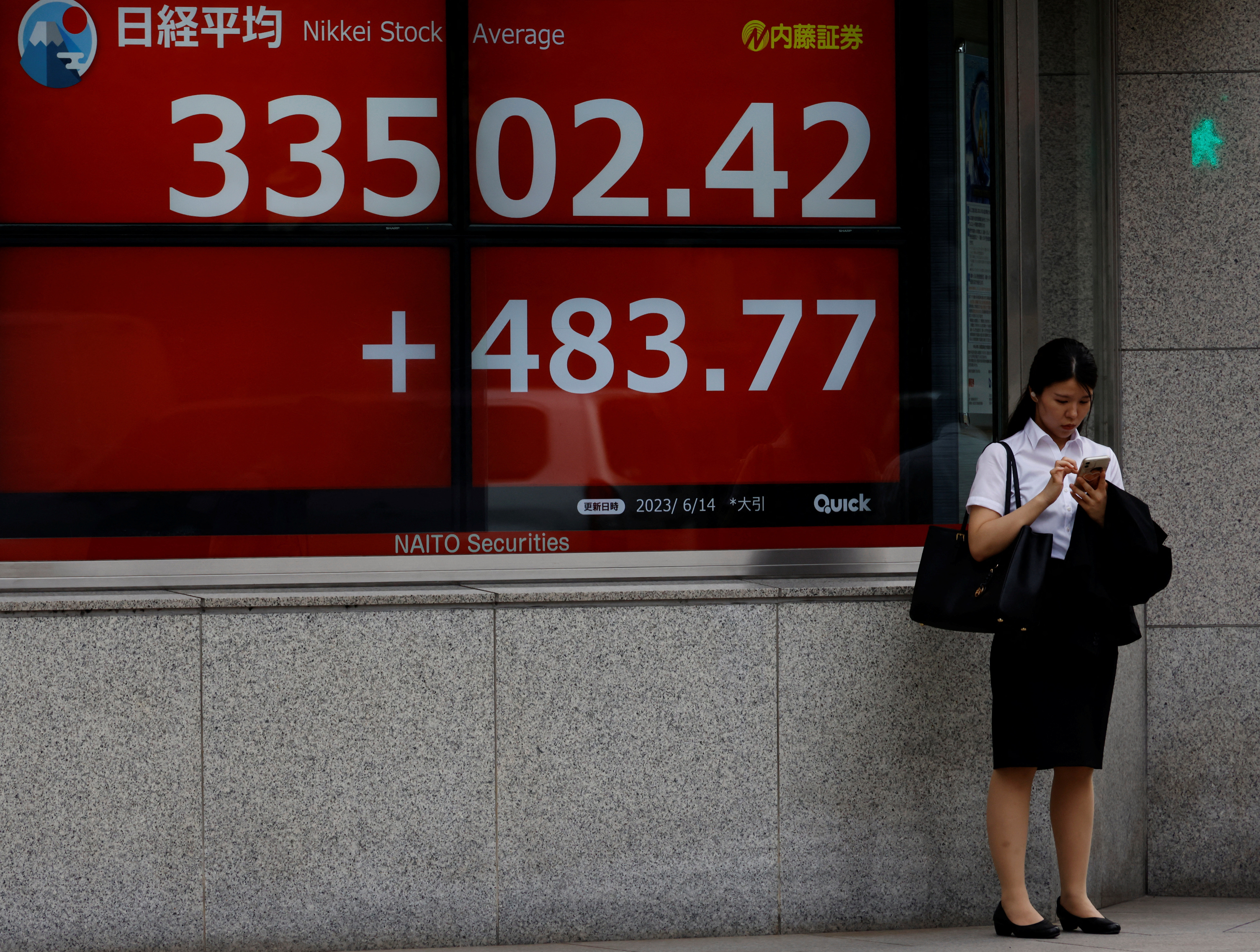 A woman uses a mobile phone in front of an electric board displaying the Nikkei stock average outside a brokerage in Tokyo