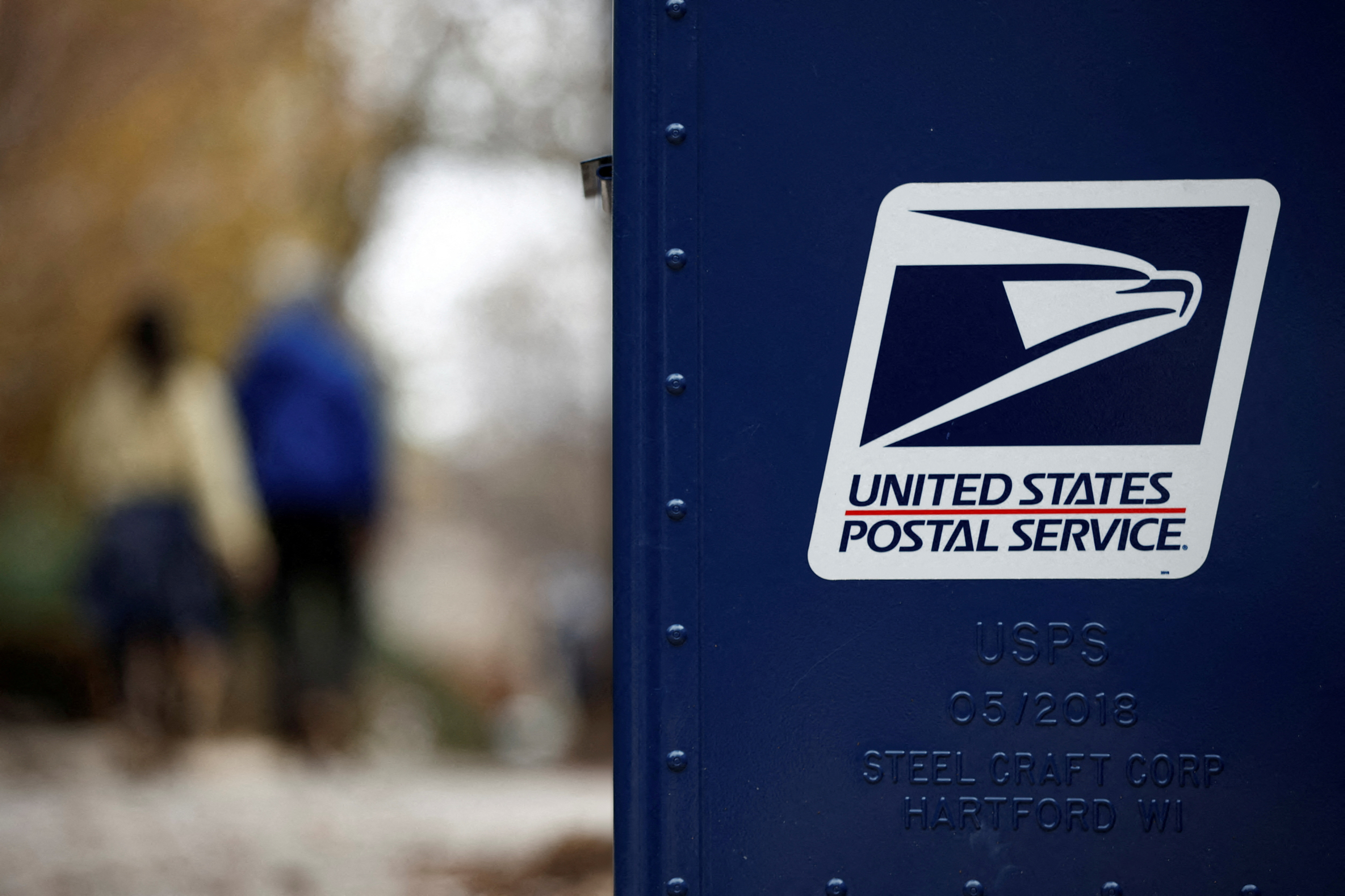 Blue USPS mail collection box with American flag emblem on side, located in a city street scene.