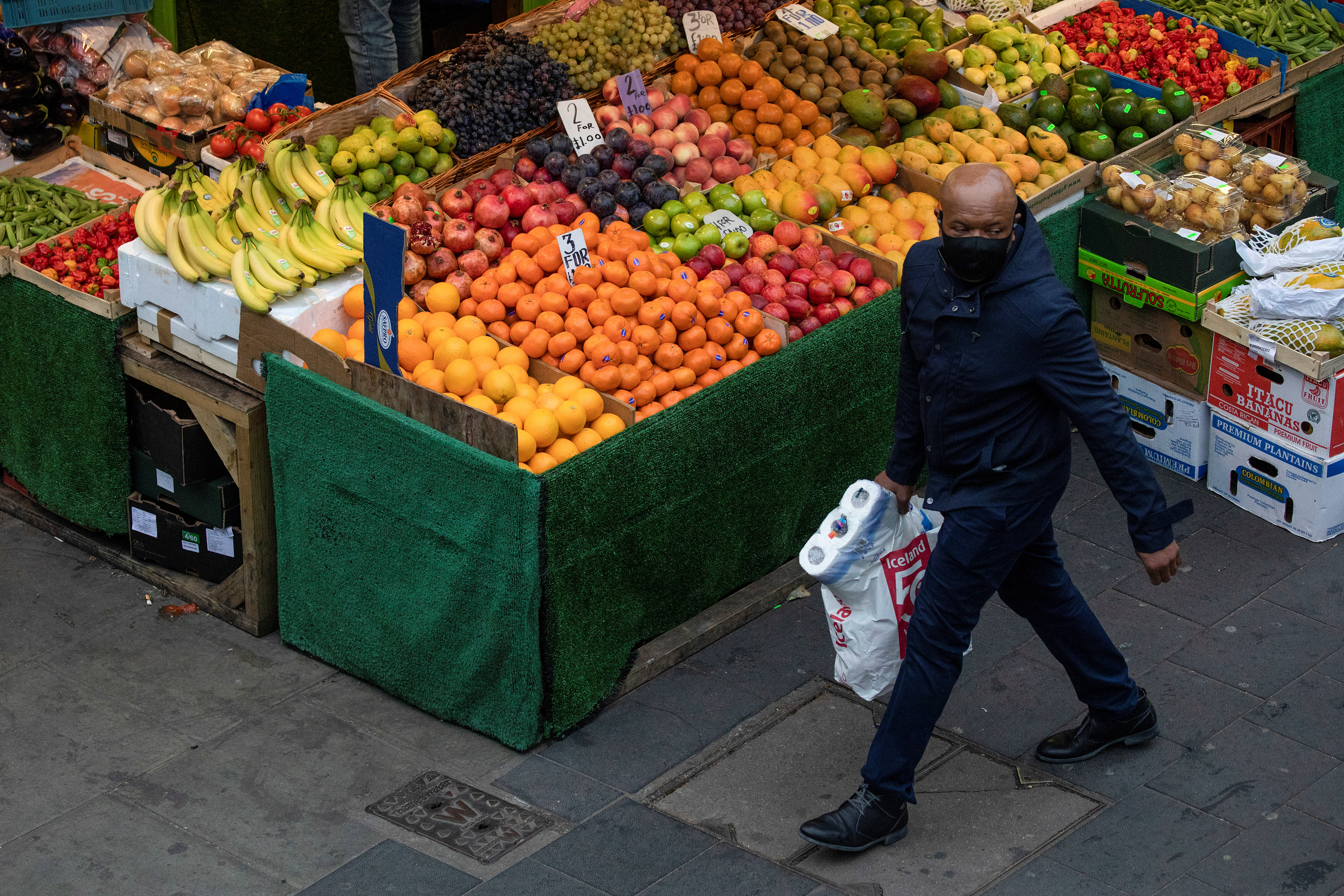 A man carrying toilet roll walks past a fruit and vegetable shop at Brixton Market, London, Britain