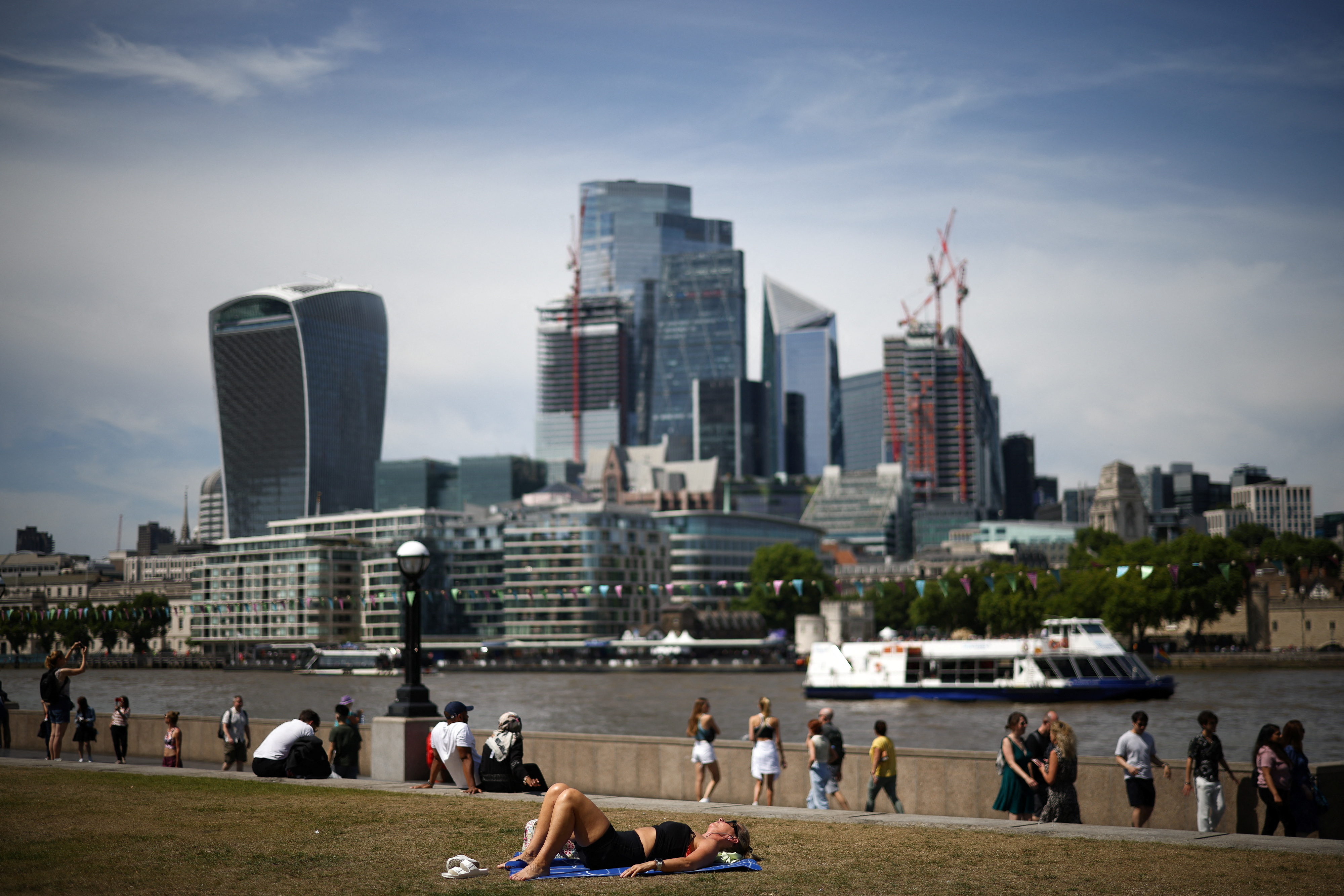 People relax in the sun of the bank the River Thames, with the The City of London financial district in the distance