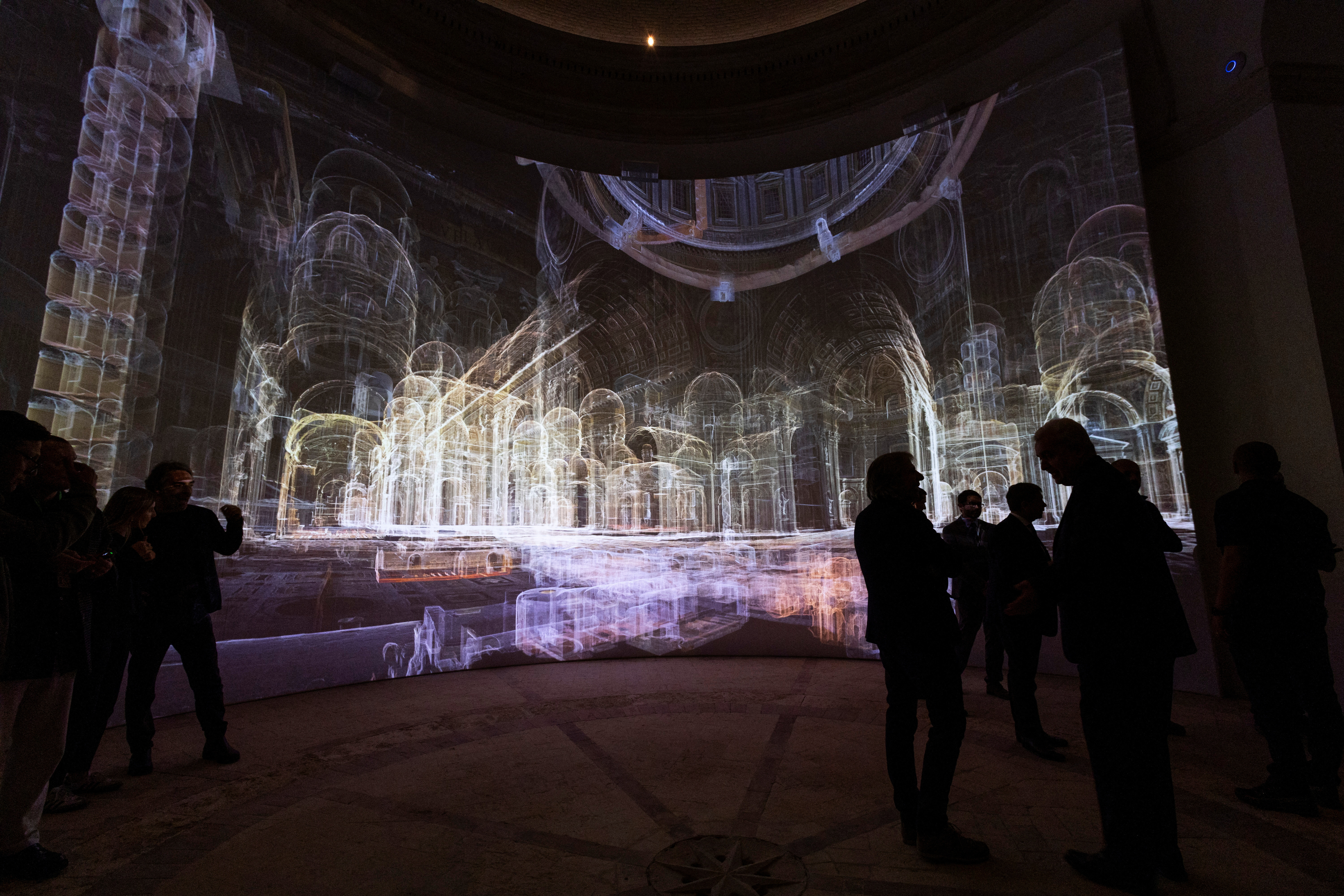 Personas observan una proyección digital de la Basílica de San Pedro en una sala oscura. La imagen proyectada muestra un modelo en 3D con líneas y contornos brillantes de la arquitectura interna de la basílica, destacando las columnas, arcos y cúpulas en tonos de luz blanca y dorada. La atmósfera es envolvente y futurista, con los espectadores en silueta contrastando con la detallada representación digital del espacio arquitectónico.