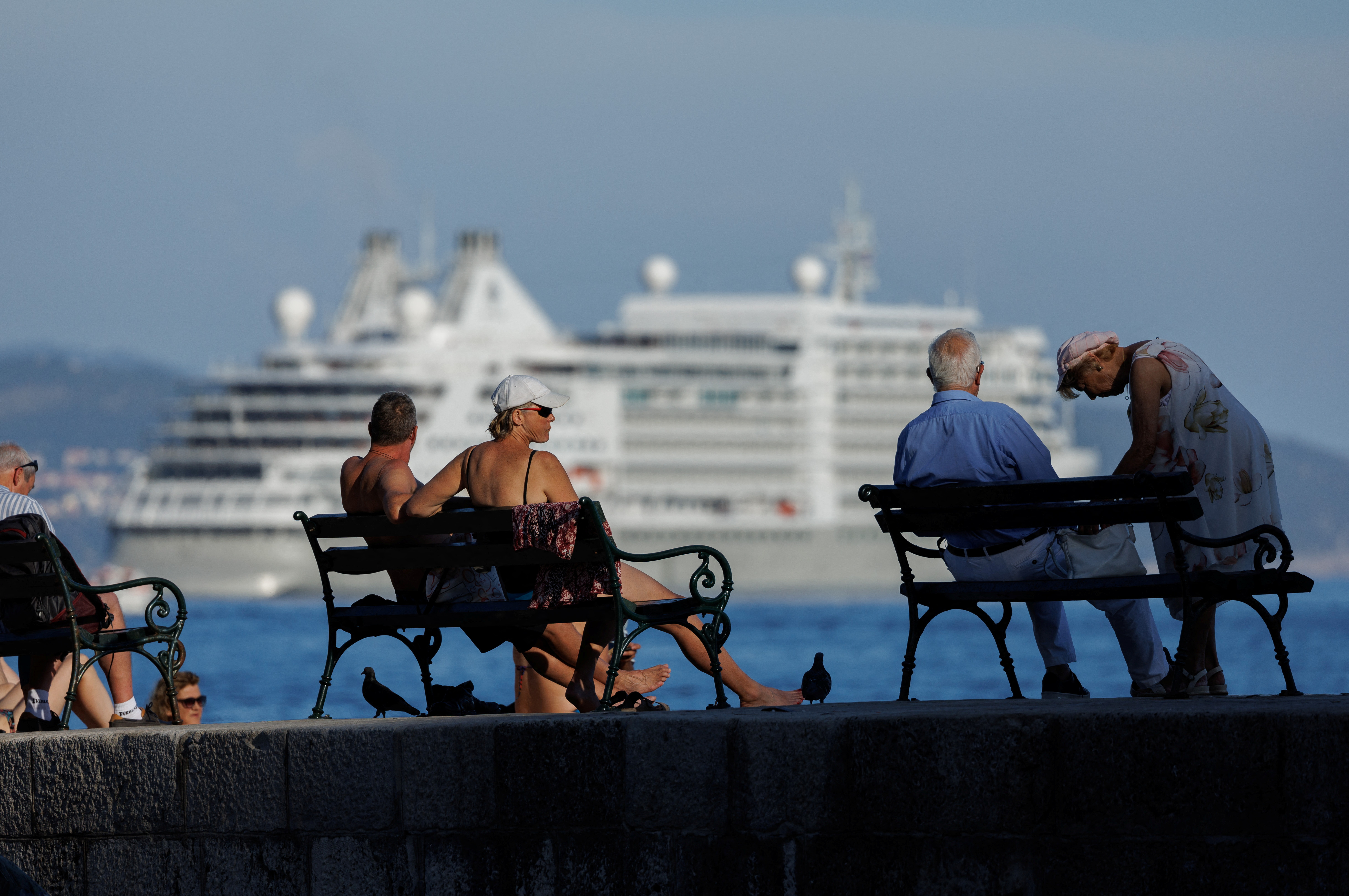 A cruise ship passes tourists resting on benches in Dubrovnik, Croatia