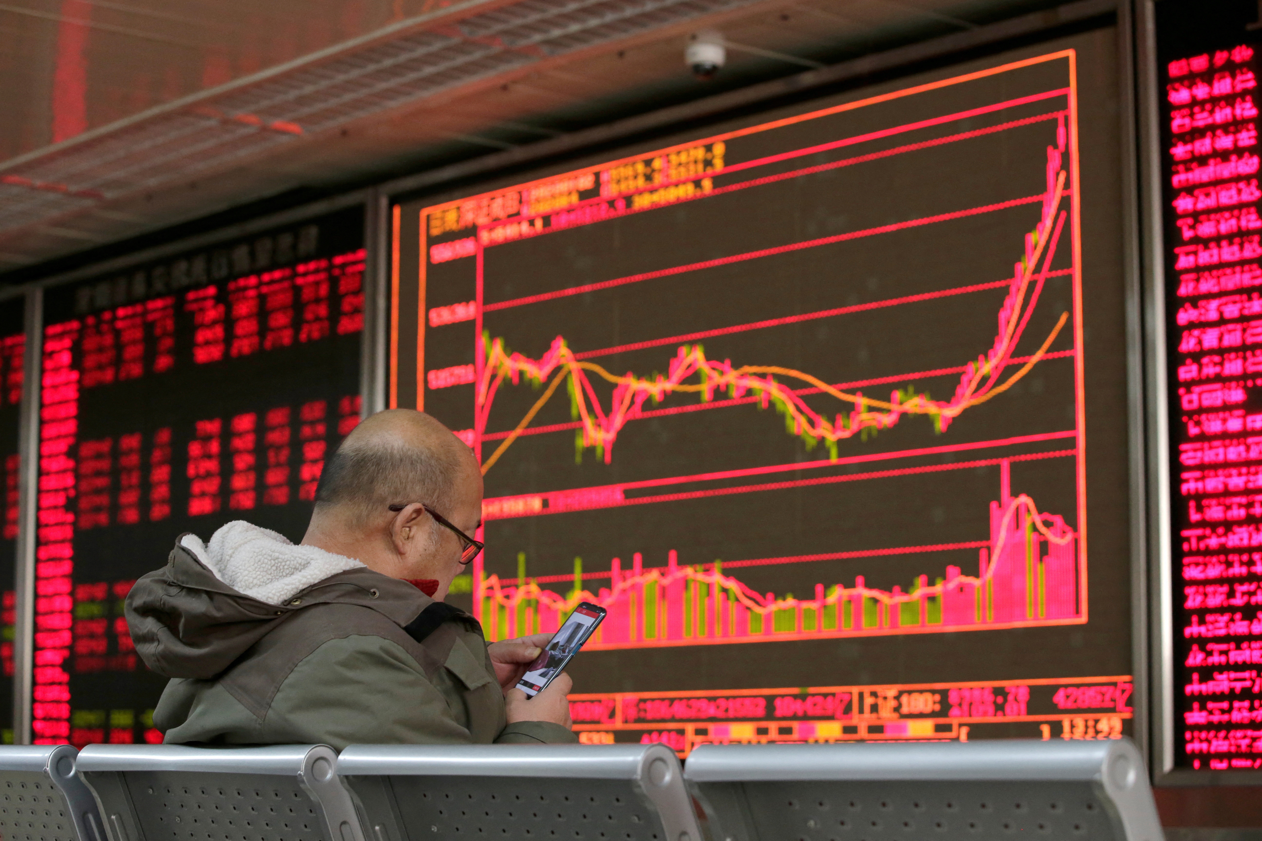 An investor looks at his mobile phone in front of a board showing stock information at a brokerage office in Beijing