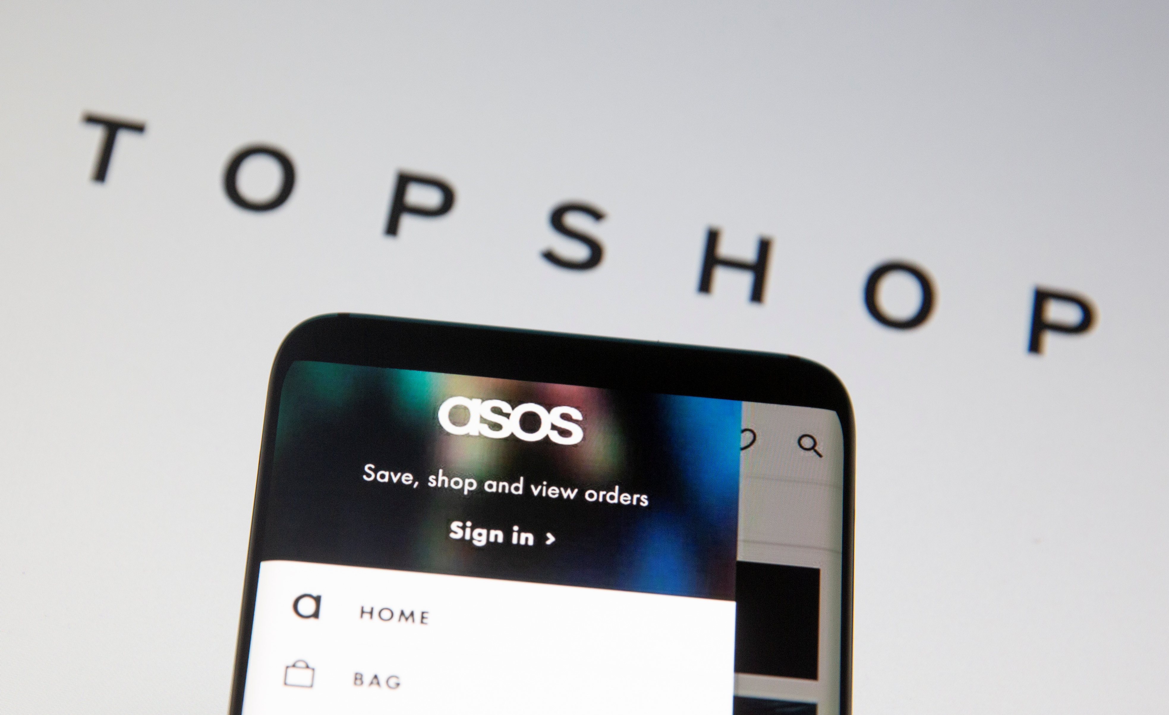 Asos logo is seen in a smartphone in front of a displayed TopShop logo in this illustration