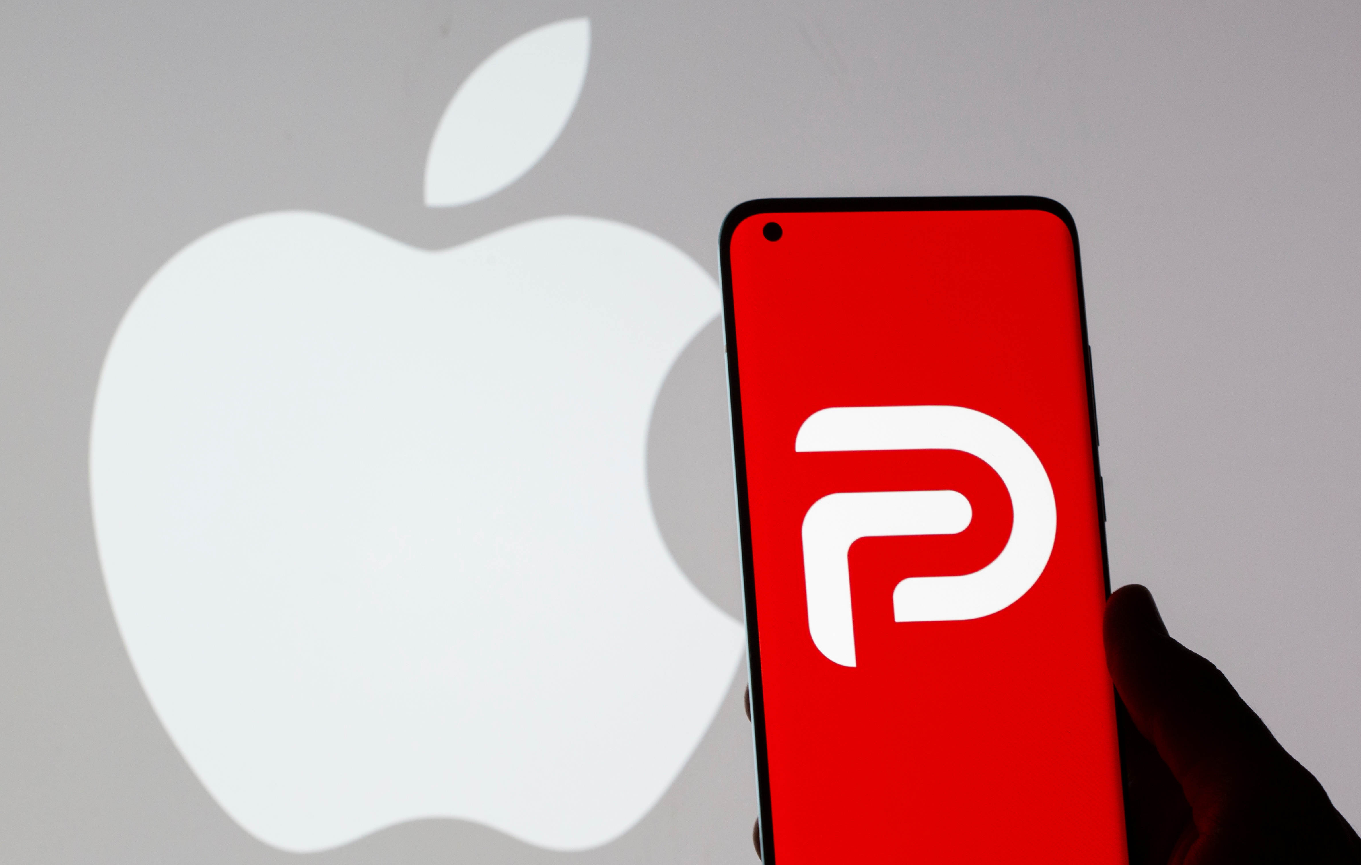 Woman holding smartphone with Parler logo in front of displayed Apple logo in this illustration