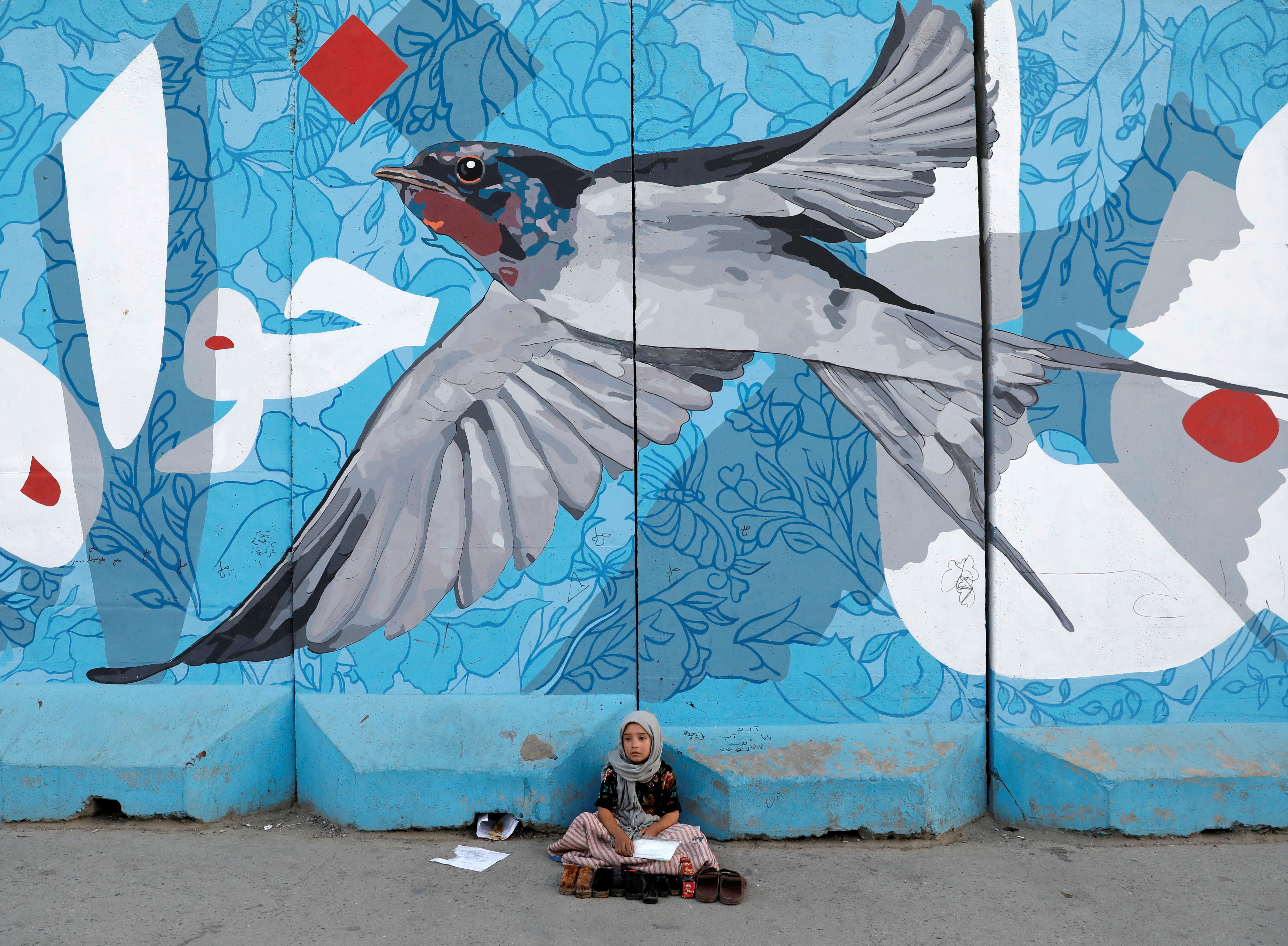 A shoe polishing girl waits for customers under graffiti on a wall in Kabul