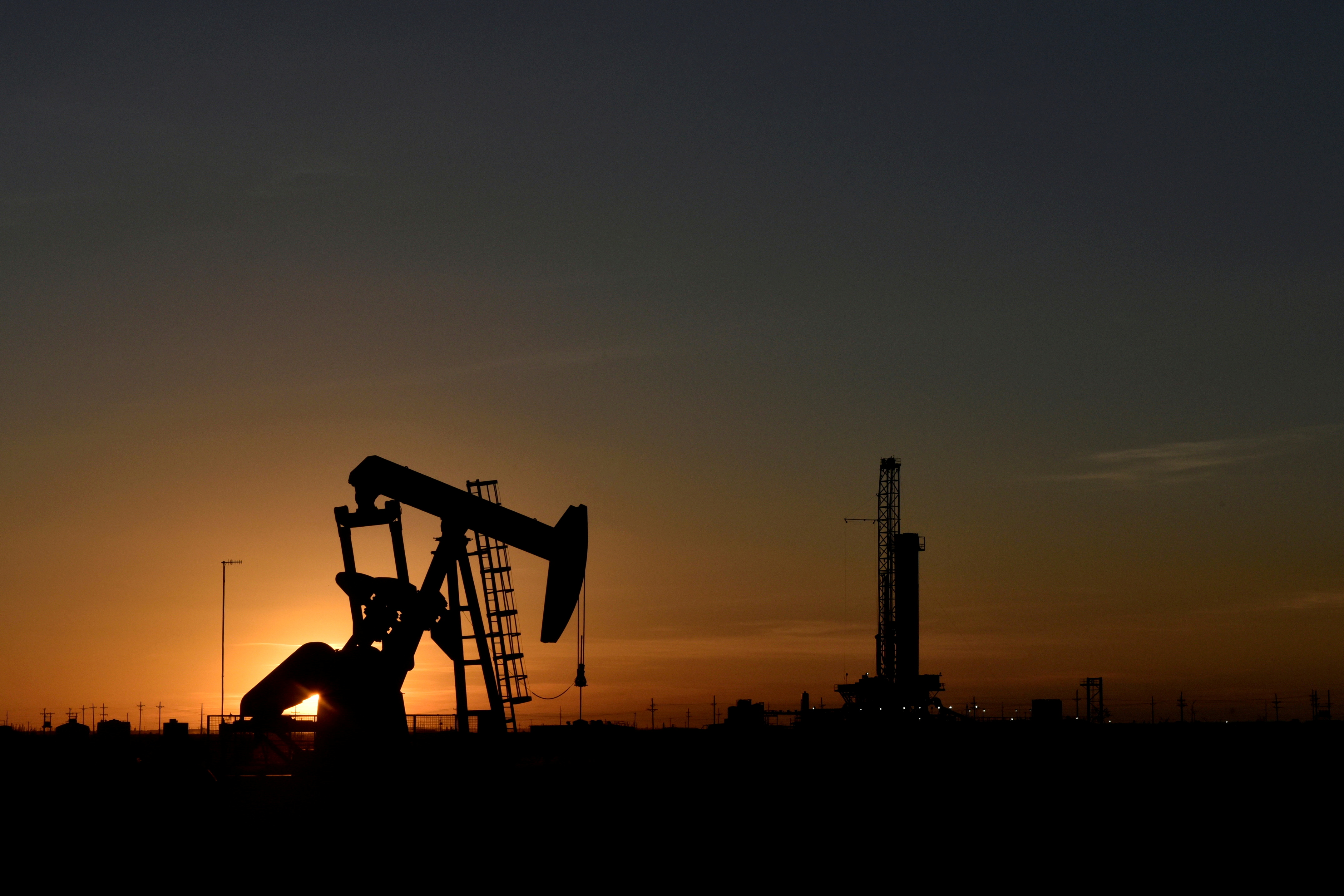 A pump jack operates in front of a drilling rig at sunset in an oil field in Midland, Texas, U.S.