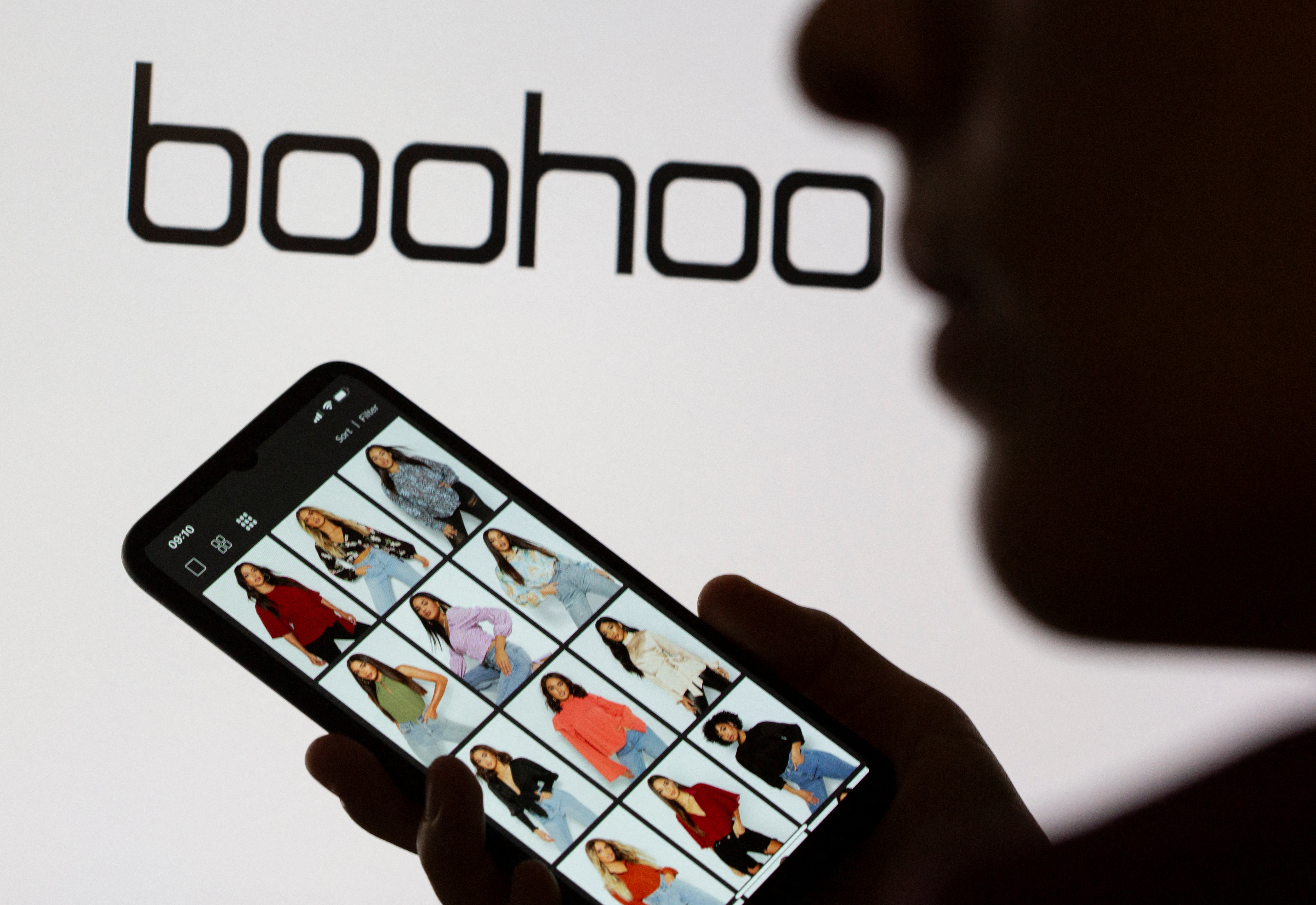 A woman poses with a smartphone showing the Boohoo app in front of the Boohoo logo on display in this illustration
