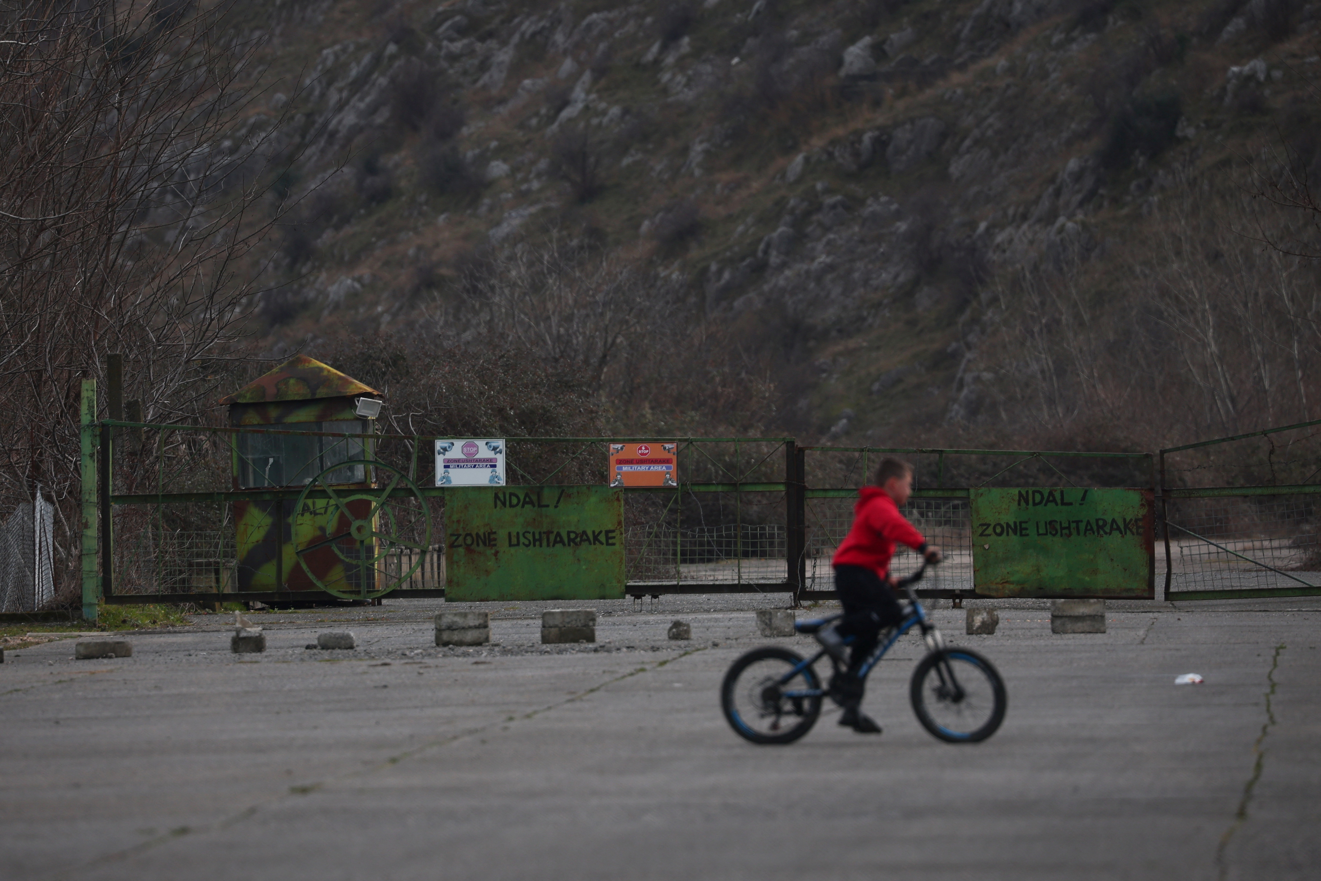 A child rides a bicycle next to a checkpoint at a military base, in Gjader emigrantë