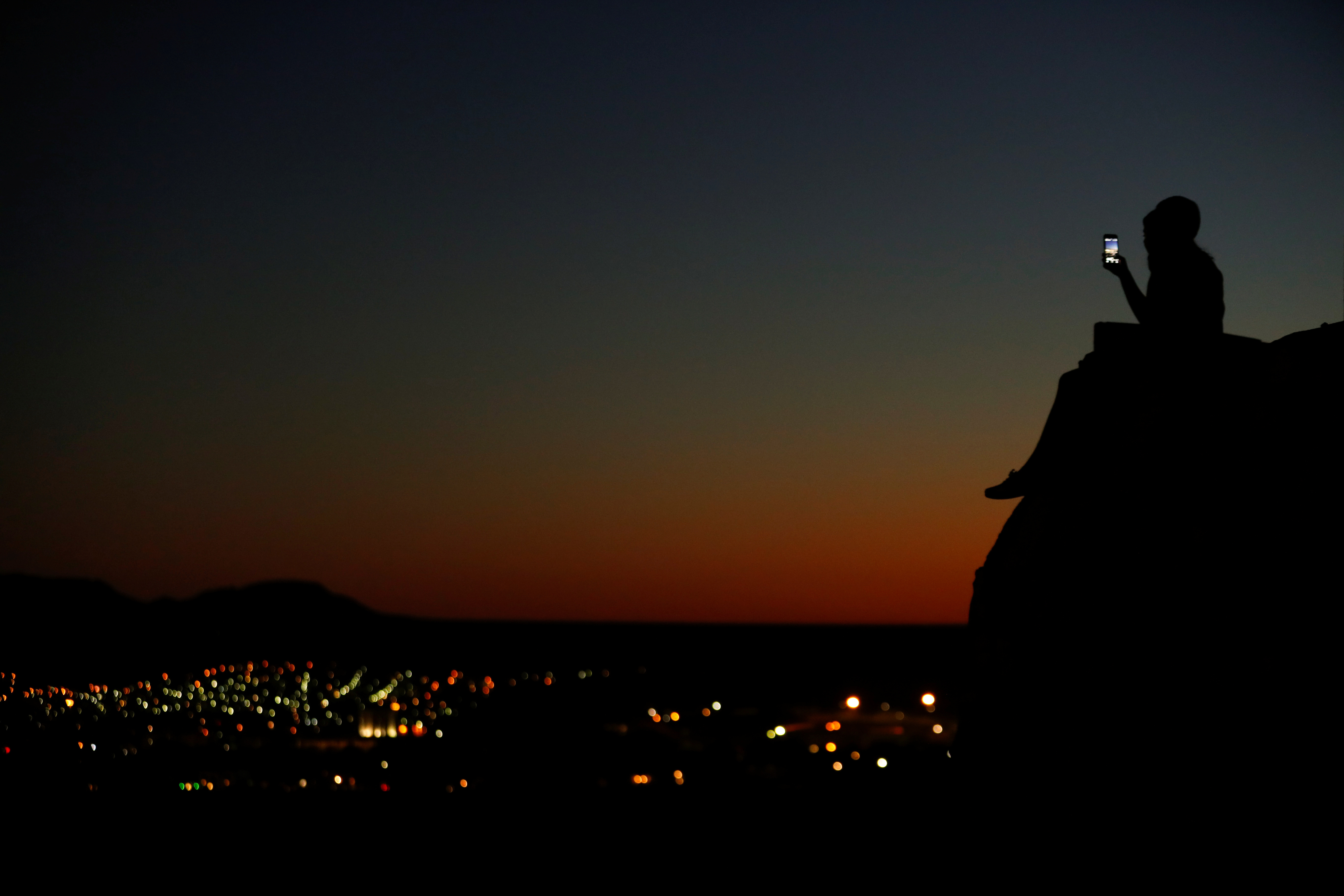 A young couple look at their phone as they sit on a hillside after sun set in El Paso, Texas
